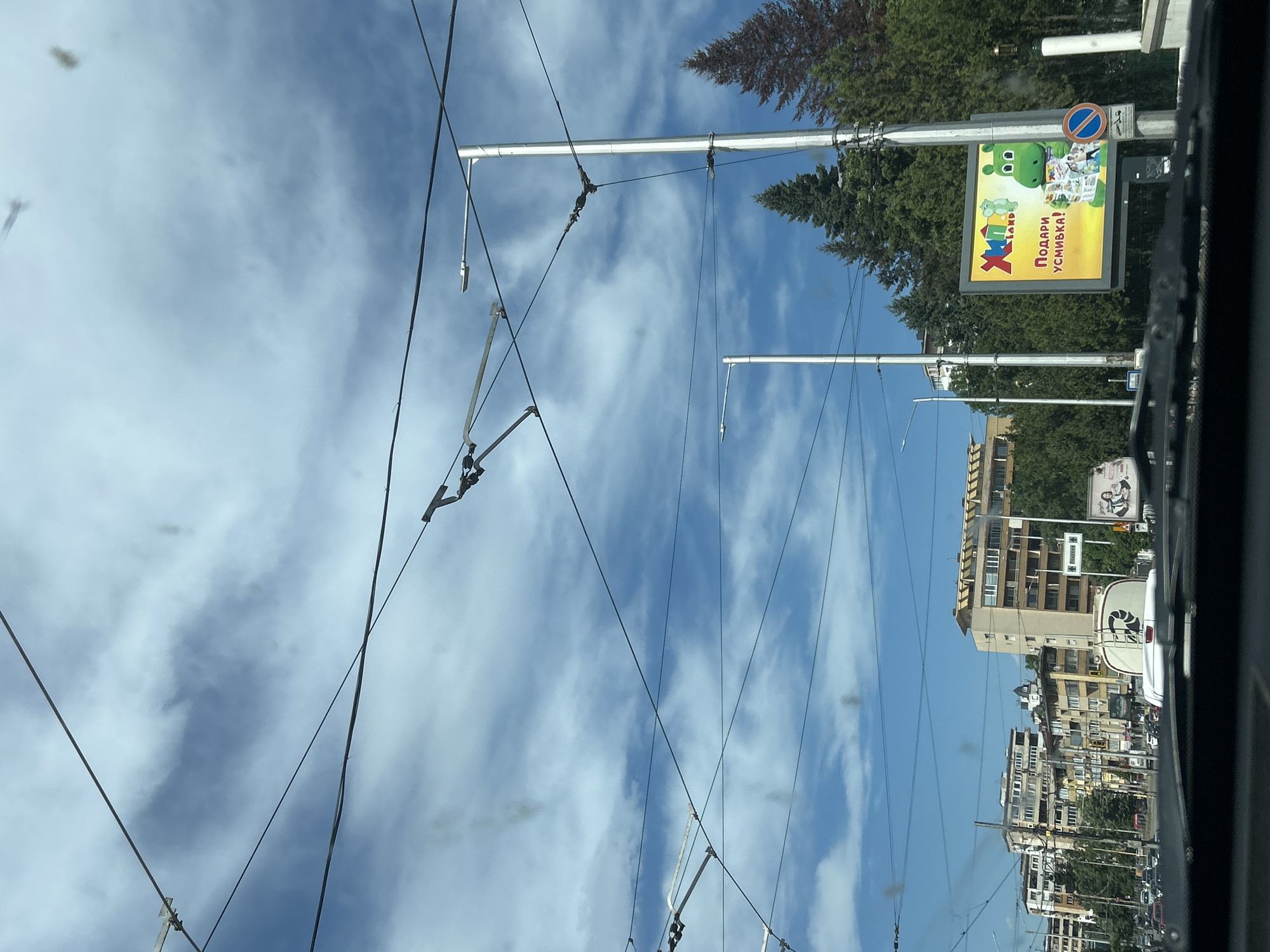The image, taken from within a vehicle, presents a view of the city of Sofia, Bulgaria. Overhead tram lines crisscross against a backdrop of a blue sky with scattered clouds. Buildings of varying heights line the street. Visible on the right side of the frame is a billboard advertising "X-Land" in Bulgarian. Trees are interspersed among the buildings and along the street. Other vehicles can be seen on the road.