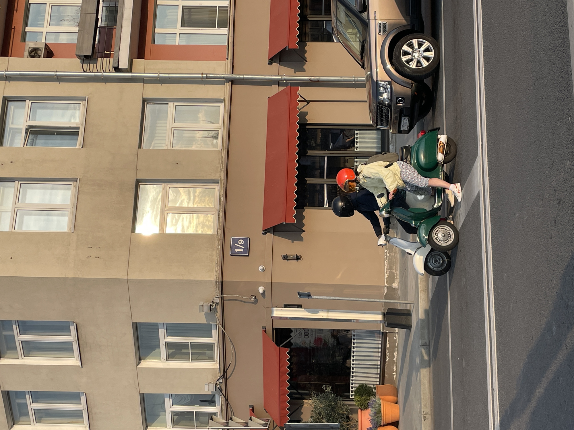The image captures a street scene in Moscow, Russia, adjacent to a building located at address 1/9. The building is multi-storied with a beige facade, visible windows, and red awnings over some of the ground floor storefronts. A bronze-colored Land Rover SUV is parked along the curb. Two individuals are interacting near a vintage green Vespa scooter. One person is wearing a black jacket, helmet, and white shoes, while the other is wearing a light-colored jacket, patterned shorts, a red helmet, and white shoes. The scene is lit with strong sunlight, casting shadows.