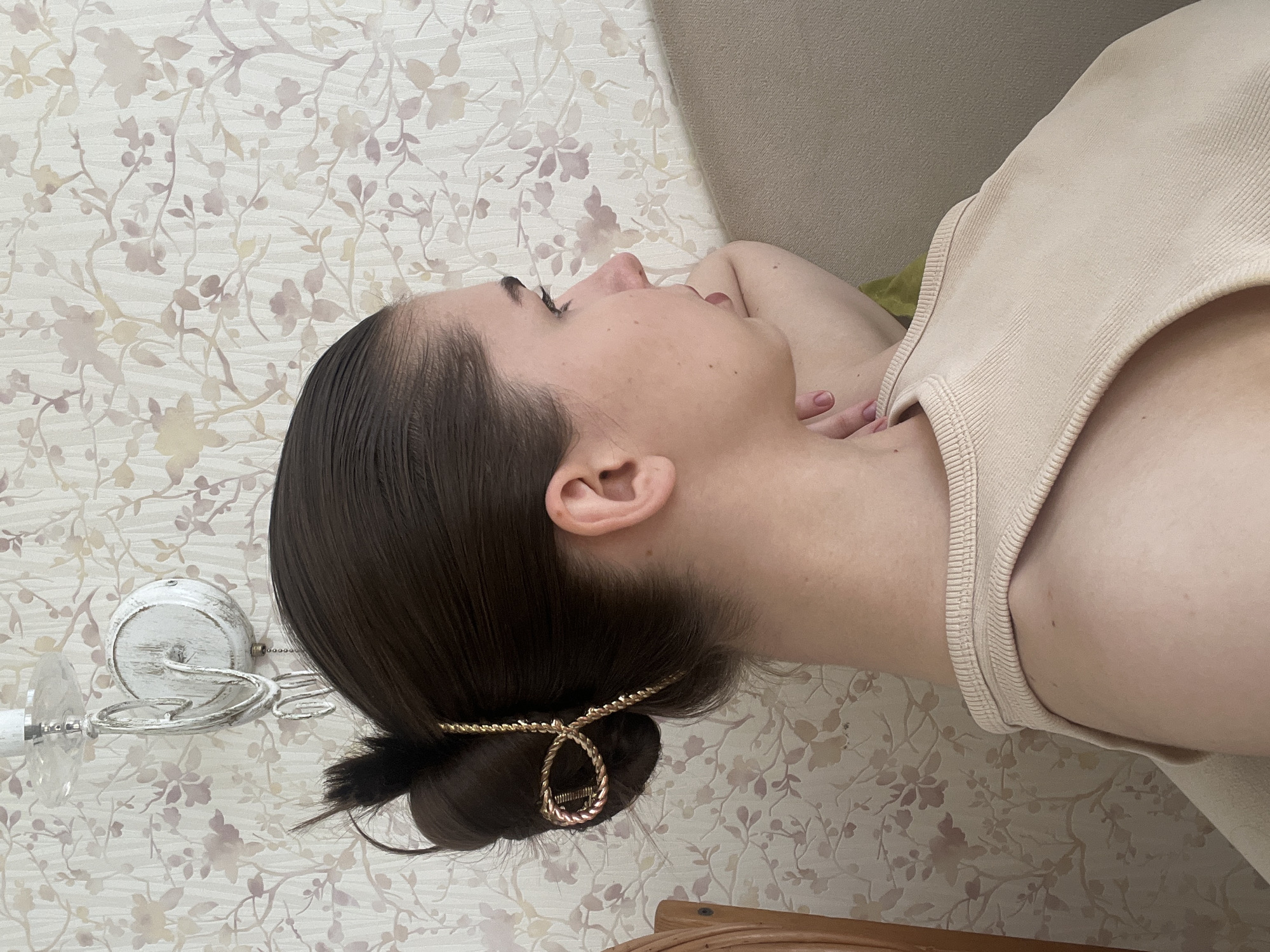A light-skinned woman lies on her back with her head turned to the left. She has dark brown hair pulled back into a bun with a gold-colored hair accessory. She wears a beige tank top. The background features a floral-patterned wallpaper. A white light fixture is visible in the background. The location is not discernible.