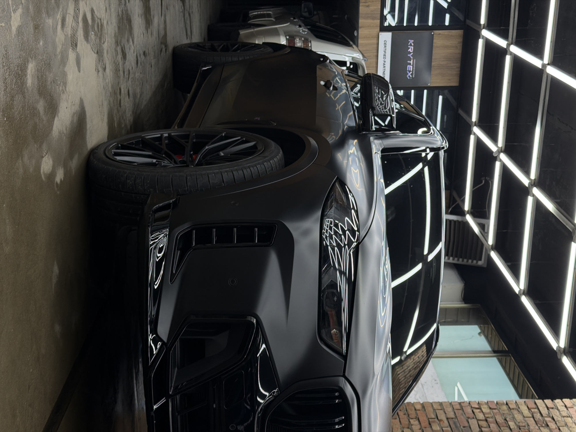 The image depicts a car detailing shop. A matte black car, likely a BMW due to the kidney grille shape, is the main subject, positioned in the foreground. Another car, appearing white, is visible partially in the background. A Krytex Certified Partner sign is mounted on a wooden wall behind the vehicles. The shop is illuminated by numerous vertical LED strip lights, creating a bright, sterile environment. The floor appears to be concrete. A brick wall and a window are visible at the bottom right corner, suggesting an exterior location. The location in the city of Krasnoyarsk, Russia, cannot be confirmed solely based on the image.