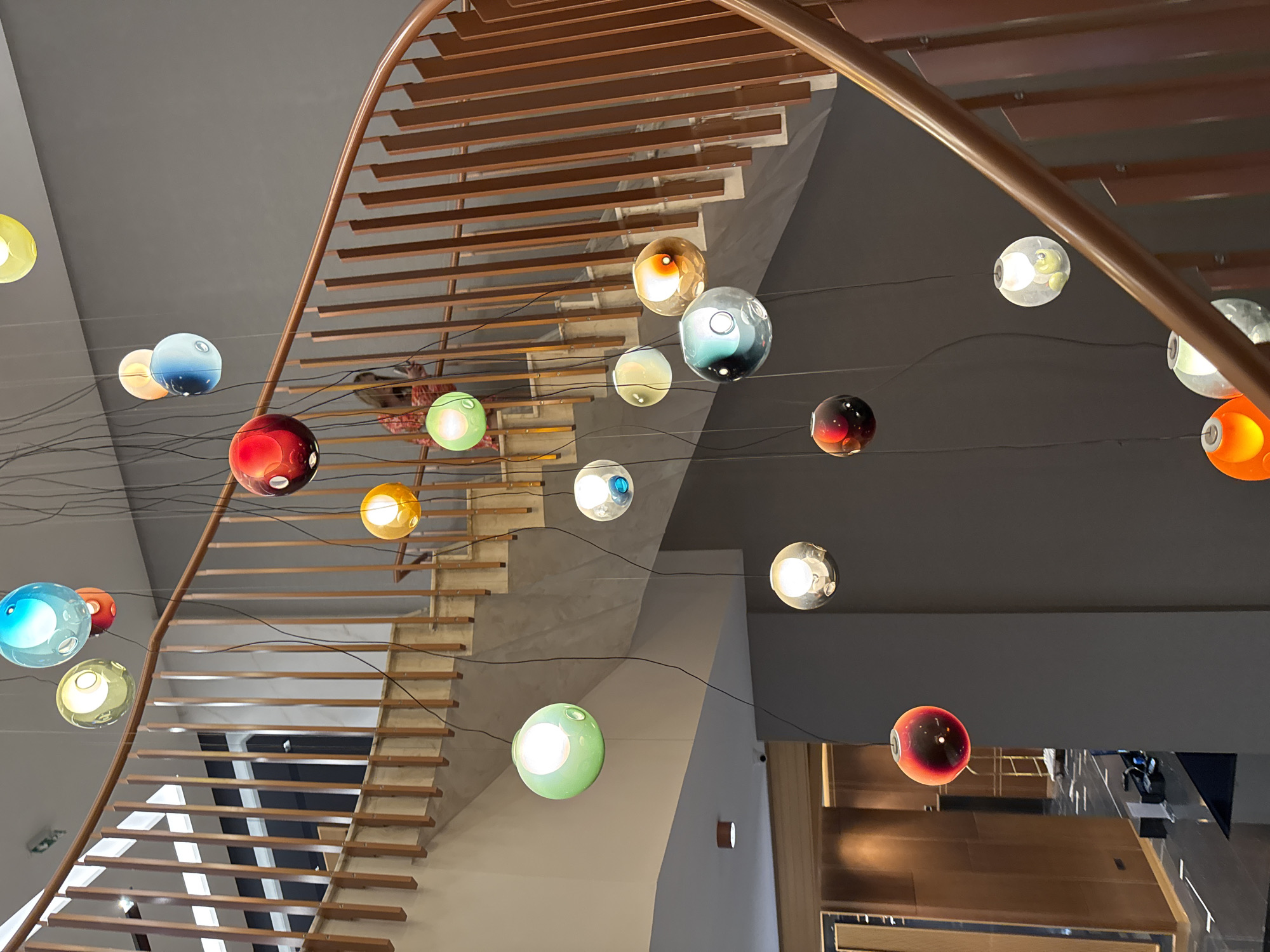 The image shows a modern interior with a curved wooden staircase and a striking light fixture composed of numerous colorful glass orbs suspended at varying heights. The staircase features light-colored risers and wooden treads with an open design, allowing light to filter through. A curved wooden handrail follows the staircase's contour. The background appears to be a neutrally colored wall, and a partial view of a hallway or entryway is visible in the lower right corner. The scene likely represents a hotel or upscale building interior, potentially in Varna, Bulgaria, though specific Varna landmarks are not visible. There are no identifiable people in the image. The focus is on the architectural design and lighting.