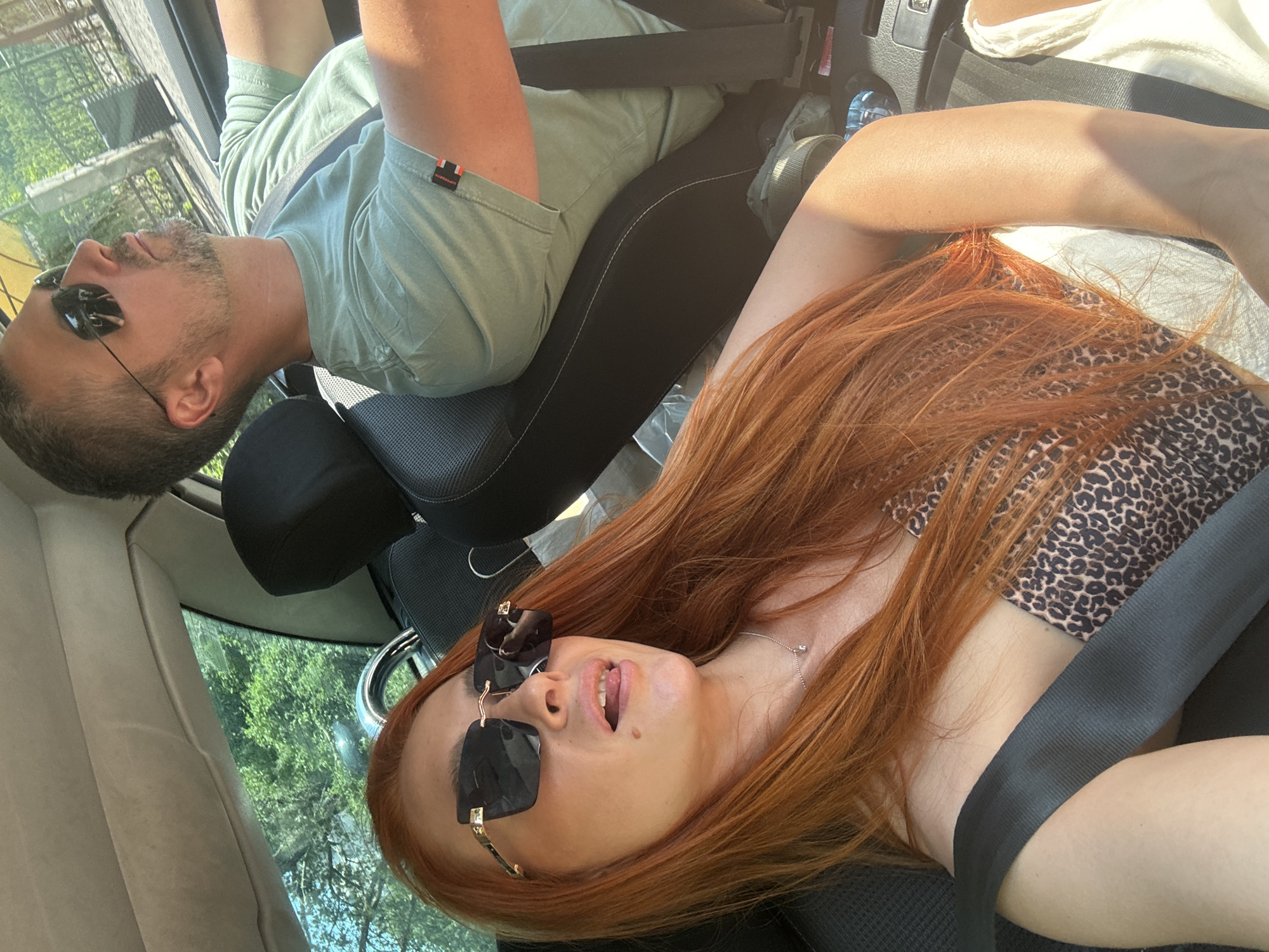 The image shows two people, a man and a woman, inside a car. The man is in the foreground, leaning back in his seat with his eyes closed and wearing sunglasses. He has short brown hair and a beard. He is wearing a light green t-shirt. The woman is in the background, also leaning back. She has long red hair and is wearing sunglasses and a leopard print top. Her mouth is slightly open. Both are wearing seatbelts. The scene appears to be during the daytime, with sunlight filtering into the car, suggesting fair weather. The background outside the car windows shows green trees and foliage, indicating an outdoor setting, possibly a rural or scenic area. The image is oriented sideways.