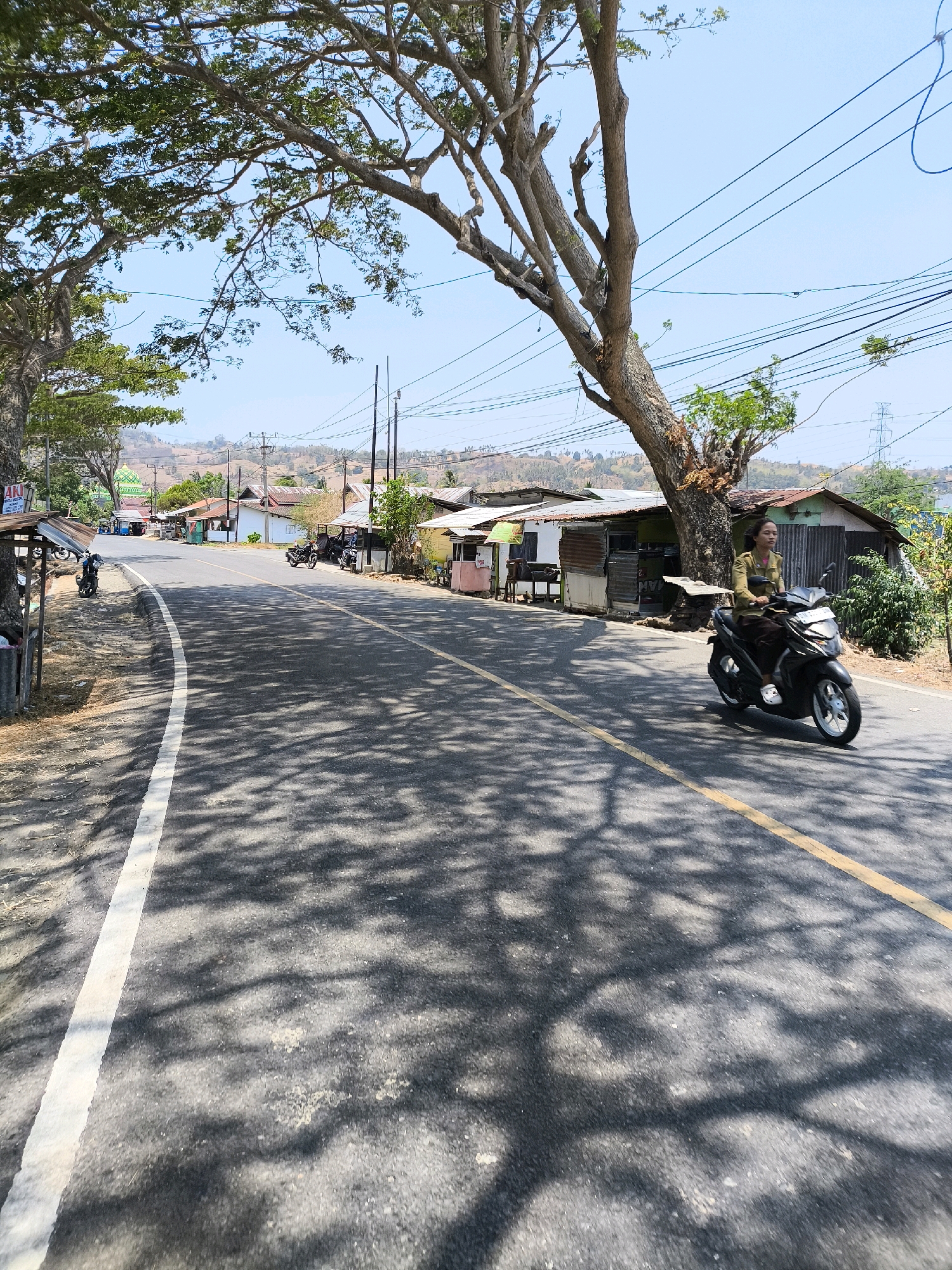 A woman rides a black scooter down a paved road lined with small buildings in Kwandang, Indonesia. The road has a white line on the left and a yellow line in the center. The woman is wearing a long-sleeved olive green shirt and dark pants. She is looking forward. The buildings appear to be shops or small residences with metal and wood construction and tin roofs. Trees with sparse foliage cast dappled shadows across the road. In the background, hills are visible under a clear blue sky with some power lines crisscrossing the scene. Several other motorcycles are parked outside the buildings.