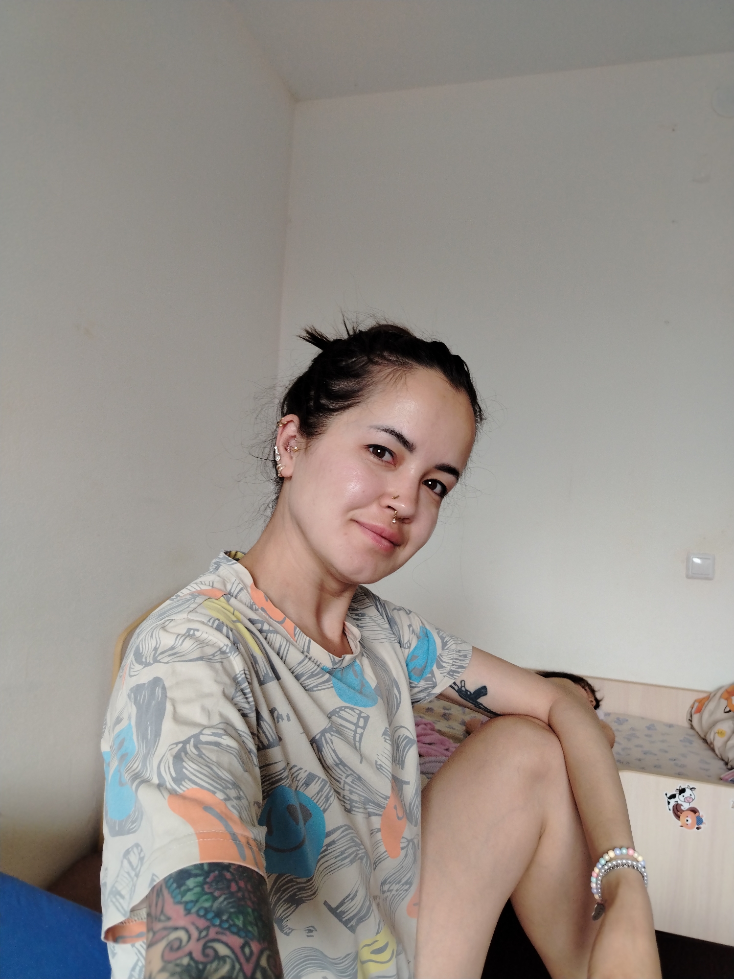 A woman sits indoors. She has brown hair pulled into a messy bun. She has multiple piercings in her left ear and a nose ring. She wears a patterned t-shirt and has tattoos on her arms. She is sitting on a blue surface in front of a bed with cartoon stickers on it. The room has white walls. The location in the city of Noviy, Russia is not determinable from the image.