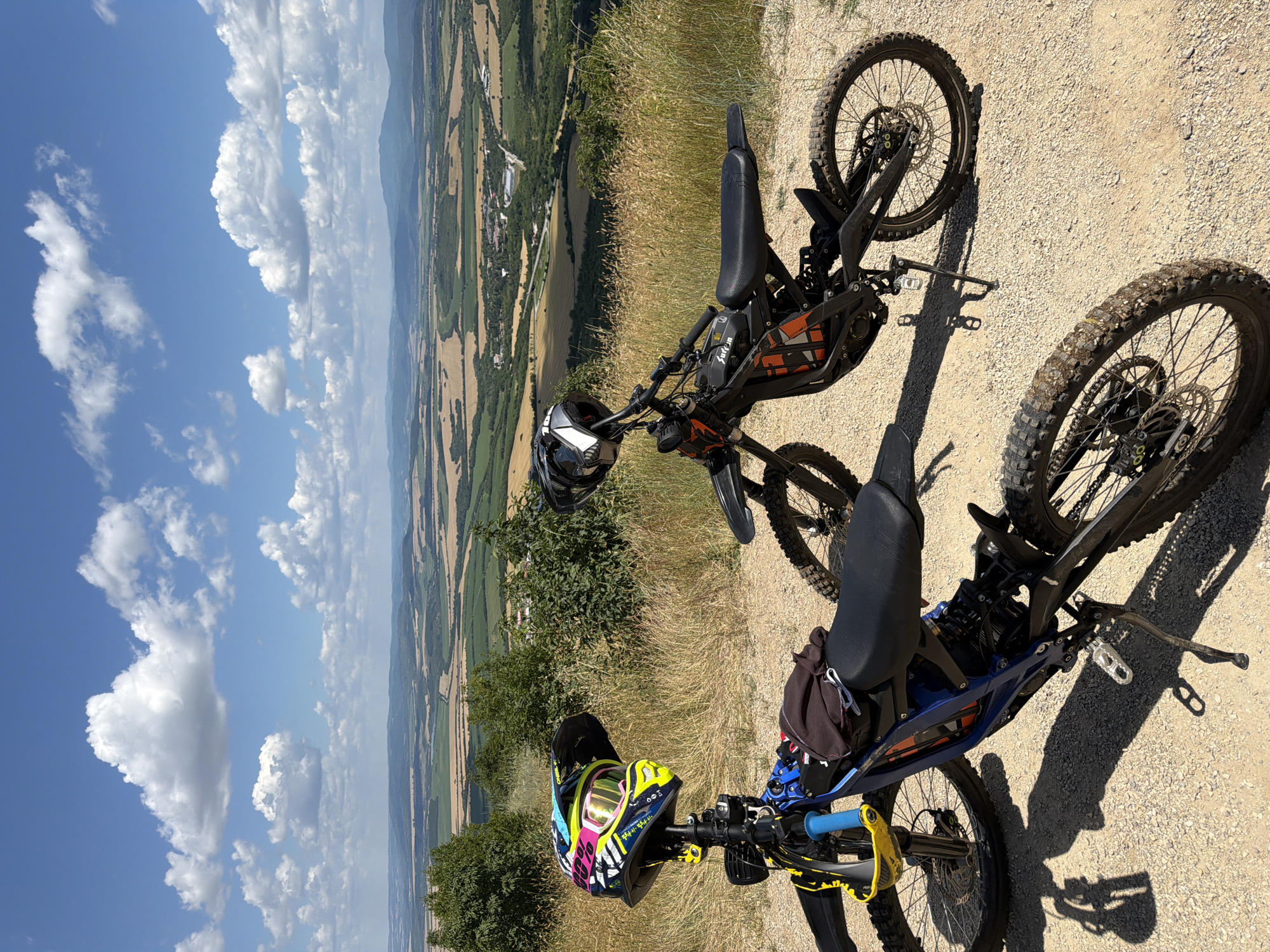 Two electric dirt bikes are parked on a gravelly path overlooking a vast, undulating landscape under a partly cloudy blue sky. The bike in the foreground is blue and black, with a colorful helmet with goggles attached to its handlebars. A dark cloth is draped over the seat. The second bike, positioned slightly behind and to the right, is black with orange accents and also has a helmet resting on its handlebars. The terrain beyond the bikes features fields of varying colors, indicating agricultural activity, interspersed with patches of green vegetation and trees. In the distance, rolling hills and mountains form the horizon. The overall impression is one of a scenic, remote location suitable for outdoor recreation. The location is specified as Sheremetia, Bulgaria.