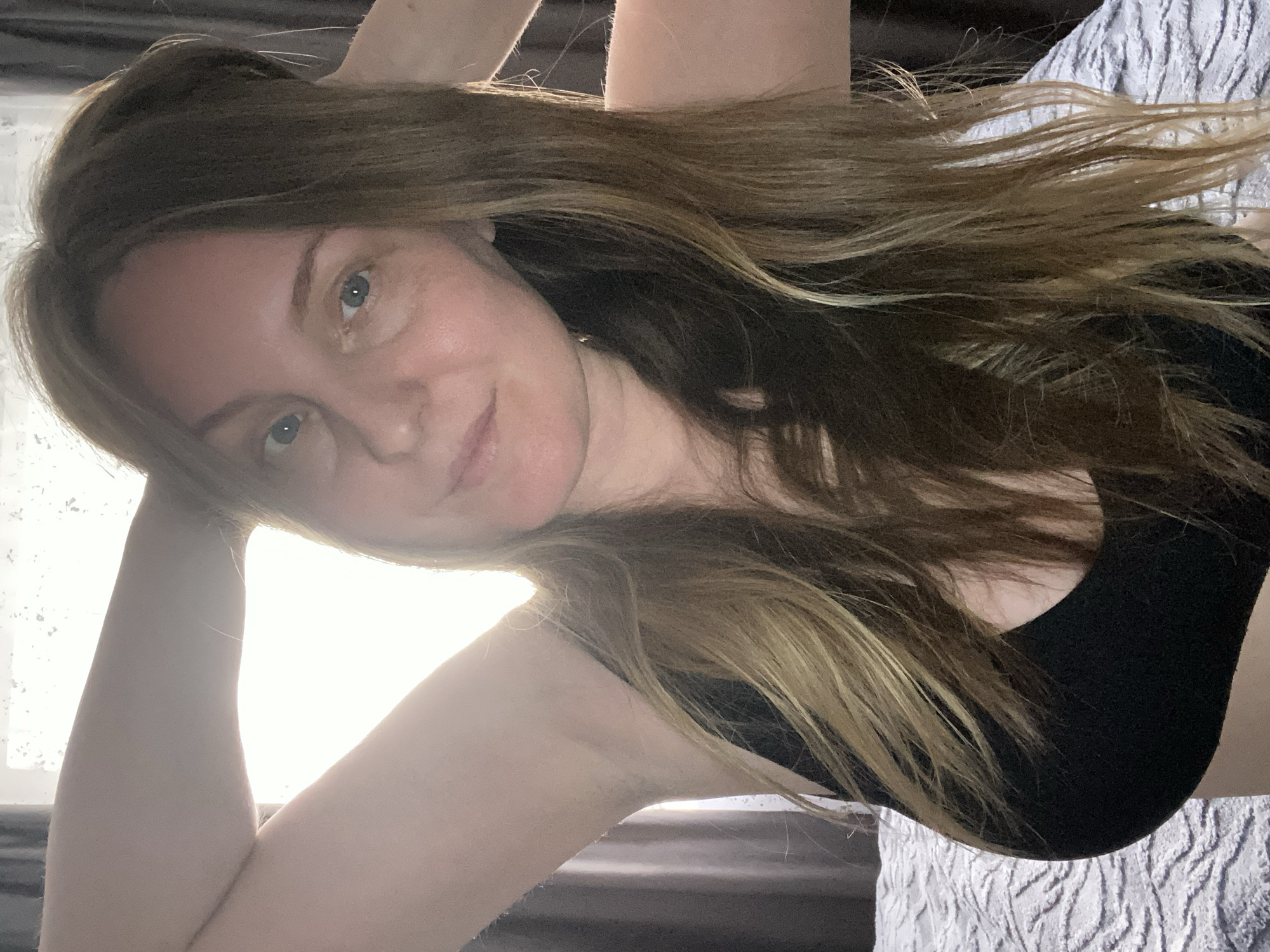 A woman with long, light brown hair and blue eyes is reclining. She is wearing a black top. Her left arm is raised behind her head, and her right arm is bent at the elbow. The background appears to be a window with bright light coming through. There are curtains on the right and bottom of the frame. The woman is resting on a white patterned surface, possibly a bed. There is no evidence to suggest the location is Noviy, Russia.