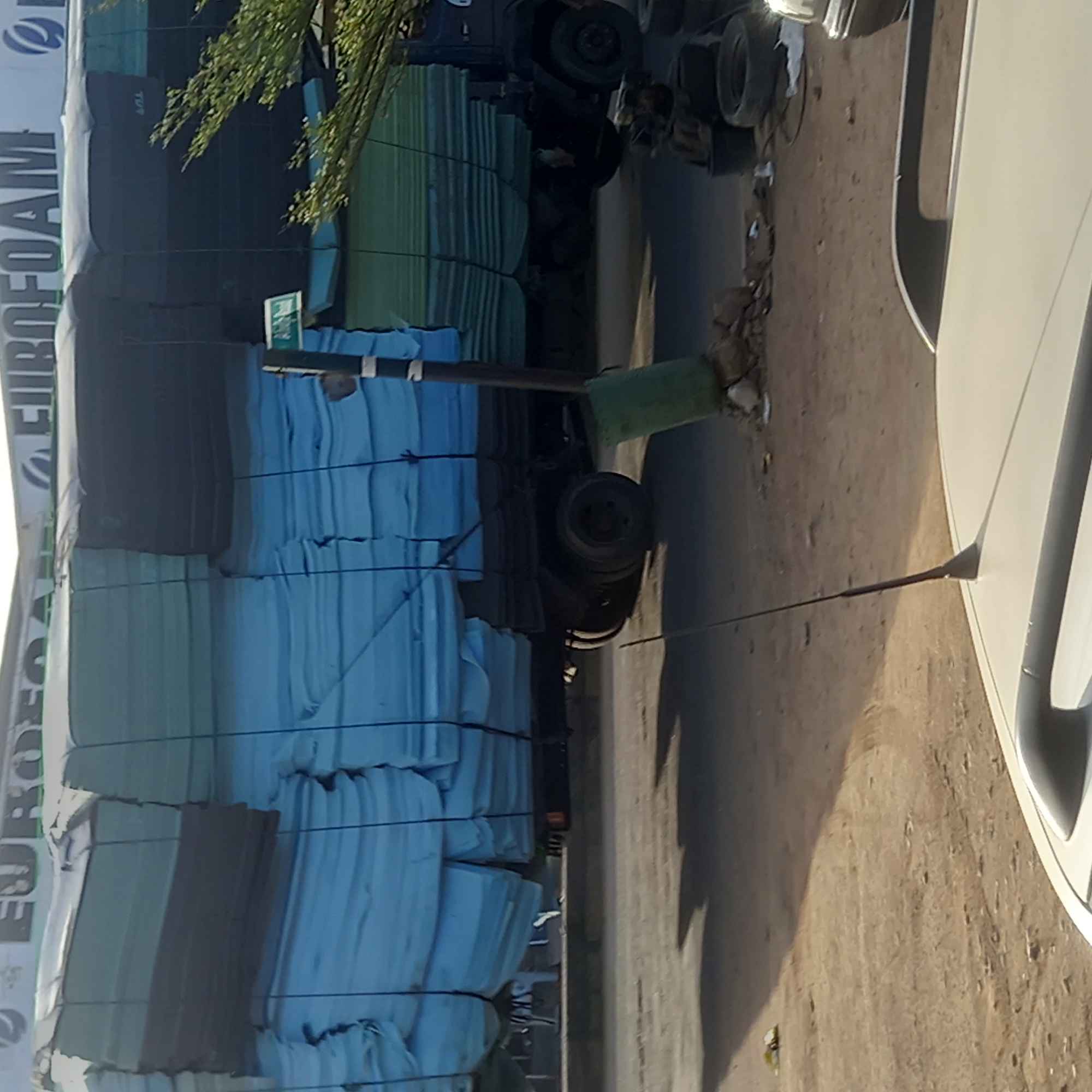 This image, taken in Dadi Riba, Nigeria, captures a large truck heavily loaded with bundles of foam products during daytime. The truck, oriented horizontally in a corrected view, occupies the left side of the frame. Its cargo consists of numerous rectangular foam sheets or mattresses, stacked high and secured with ropes or straps. Many bundles are a light blue color, while others are a darker green or grey.
Visible text on the side of the truck's load clearly spells "EUROFOAM" in large, white letters, accompanied by a blue and white logo that appears to be a stylized 'E' or wave pattern. On the right side of the image, the front quarter of a white car is partially visible, parked on what appears to be a dusty or gravelly surface.
In the mid-ground, a small green sign is mounted on a pole, with the word "MAIN" legible on it. Shadows cast by the vehicles and objects suggest bright sunlight. A tree branch with green leaves extends into the upper left of the frame. The overall scene depicts a commercial transport operation, possibly a delivery or loading area.