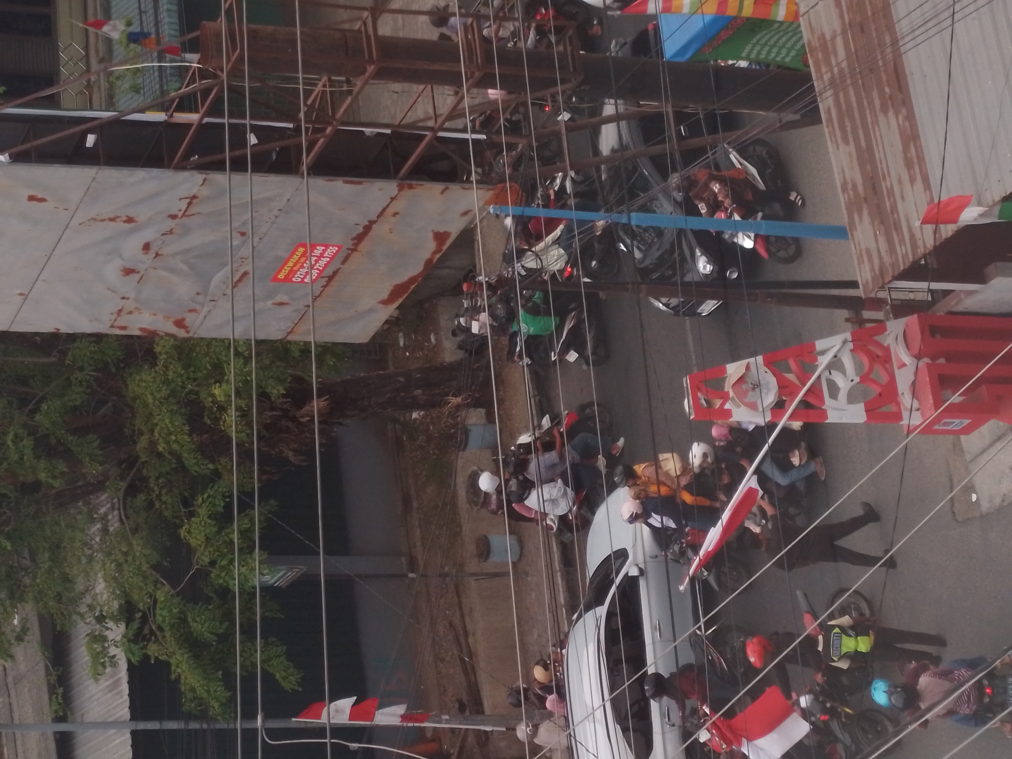 This high-angle, full-frame photograph captures a busy street scene in Bumirejo, Indonesia. The image is dominated by a collection of motorcycles and a single silver car, indicating moderate vehicular traffic. Numerous individuals are visible, mostly riding on or standing near motorcycles. Many are wearing helmets, and some are adorned with flags, suggesting a possible event or celebration.
In the foreground, a person wearing a bright yellow vest with "SECURITY" printed on it stands in the middle of the road, appearing to direct or observe traffic. Several motorcycles are clustered around this individual. To the left, a white and red Indonesian flag is prominently displayed on a pole.
Further into the scene, a variety of motorcycles, some carrying multiple passengers, navigate the asphalt road. People are dressed in casual clothing, with some wearing bright colors. The environment includes trees, electrical wires crisscrossing the frame, and buildings with corrugated metal roofs. A large, rusty metal structure, possibly a billboard or construction material, stands on the right side of the image, bearing some faded text and what appears to be an advertisement. The overall impression is one of an active, somewhat chaotic urban environment.