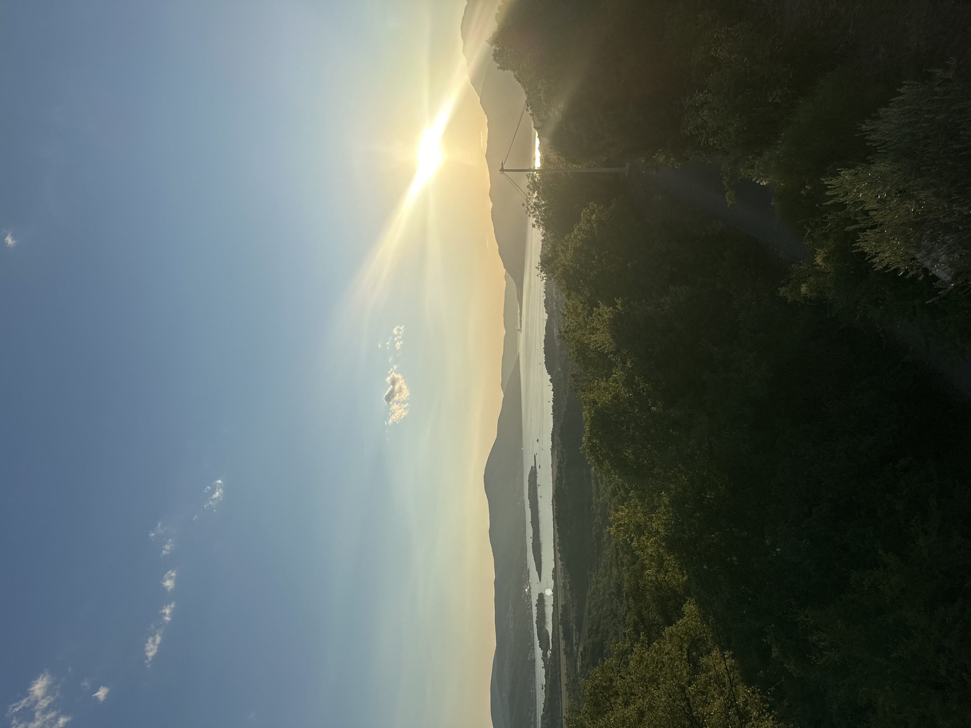 A telephoto shot captures a serene sunset over a bay in Kavač, Montenegro, with sun rays streaming through a lone cloud and casting a golden hue on the landscape. The foreground is dominated by lush green trees, with a paved path winding through them. In the mid-ground, a tranquil bay is dotted with boats, while distant hills form a majestic backdrop. The sky is a gradient of blue, transitioning to yellow and orange near the horizon, as the sun dips low.
