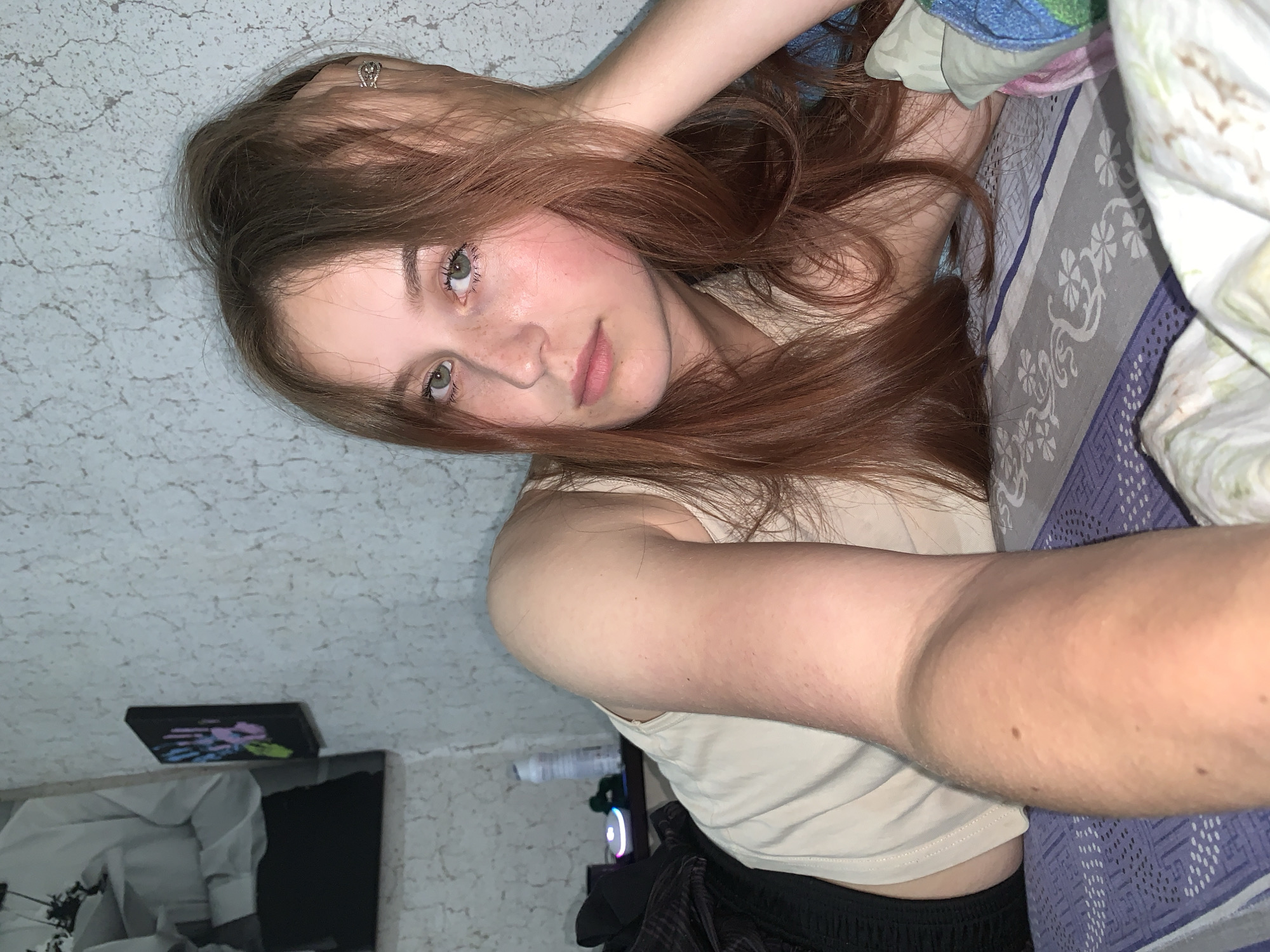 A young woman with long, reddish-brown hair and light green eyes lies on what appears to be a bed, taking a selfie or being photographed from a slightly overhead angle. Her head is tilted, and her gaze is directed towards the camera. She has a scattering of light freckles across her nose and upper cheeks. Her expression is neutral and calm.
She is wearing a light-colored, possibly beige or off-white, sleeveless top. A ring with a detailed design is visible on the ring finger of her right hand, which is raised and rests near her head, lightly touching her hair.
She is positioned next to patterned bedding, primarily a blue and purple sheet with a white floral or geometric design. Other crumpled fabrics in blue, green, and pink are visible near her head, suggesting pillows or blankets.
The background on the left features a textured wall, possibly painted a light color with a distressed finish. On the lower right, a dark object with glowing colored lights (blue, pink, green) hints at electronics like a gaming setup or computer. On the lower left, a dark rectangular device, possibly a tablet or console, with vibrant blue, pink, green, and yellow graphics on its screen, is resting on a surface. Below it, a partially visible black and white framed picture or poster can be seen.
The scene is indoors, likely a bedroom, with subdued or artificial lighting. No specific time of day or weather conditions can be determined. There is no discernible text visible in the image. While the location context is Фоки, Russia, no specific visual cues in the image confirm this.