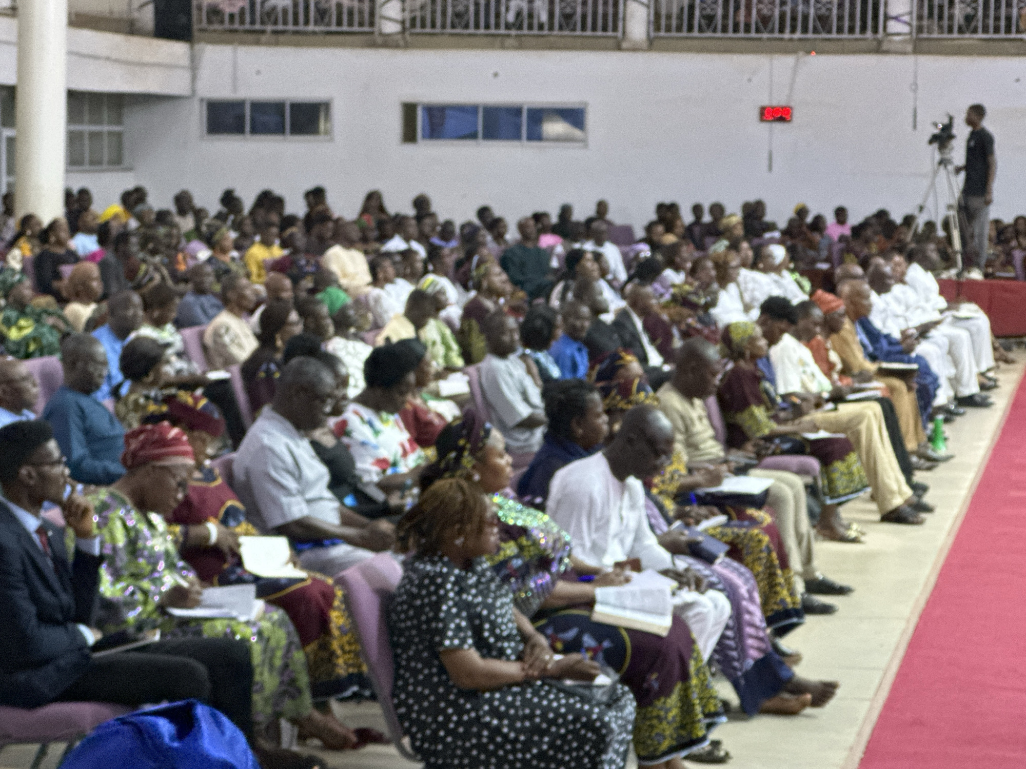 The image shows an indoor gathering of a large group of people, likely at a religious service or a public event, in Television, Nigeria. The attendees are seated in rows of chairs, facing towards the front of the venue, which is not fully visible. Most individuals are dressed in traditional Nigerian attire, characterized by vibrant colors and patterns. Some people are holding books or papers, suggesting they might be taking notes or following along with a presentation.
In the background, a white wall features a rectangular window and a digital display showing red numbers. A camera operator with a tripod is visible on the right side of the frame, positioned on a raised platform, indicating that the event is being recorded. The overall atmosphere appears to be one of attentive participation, with the majority of people looking forward, engaged in the proceedings. The lighting is somewhat dim, typical of an indoor venue.