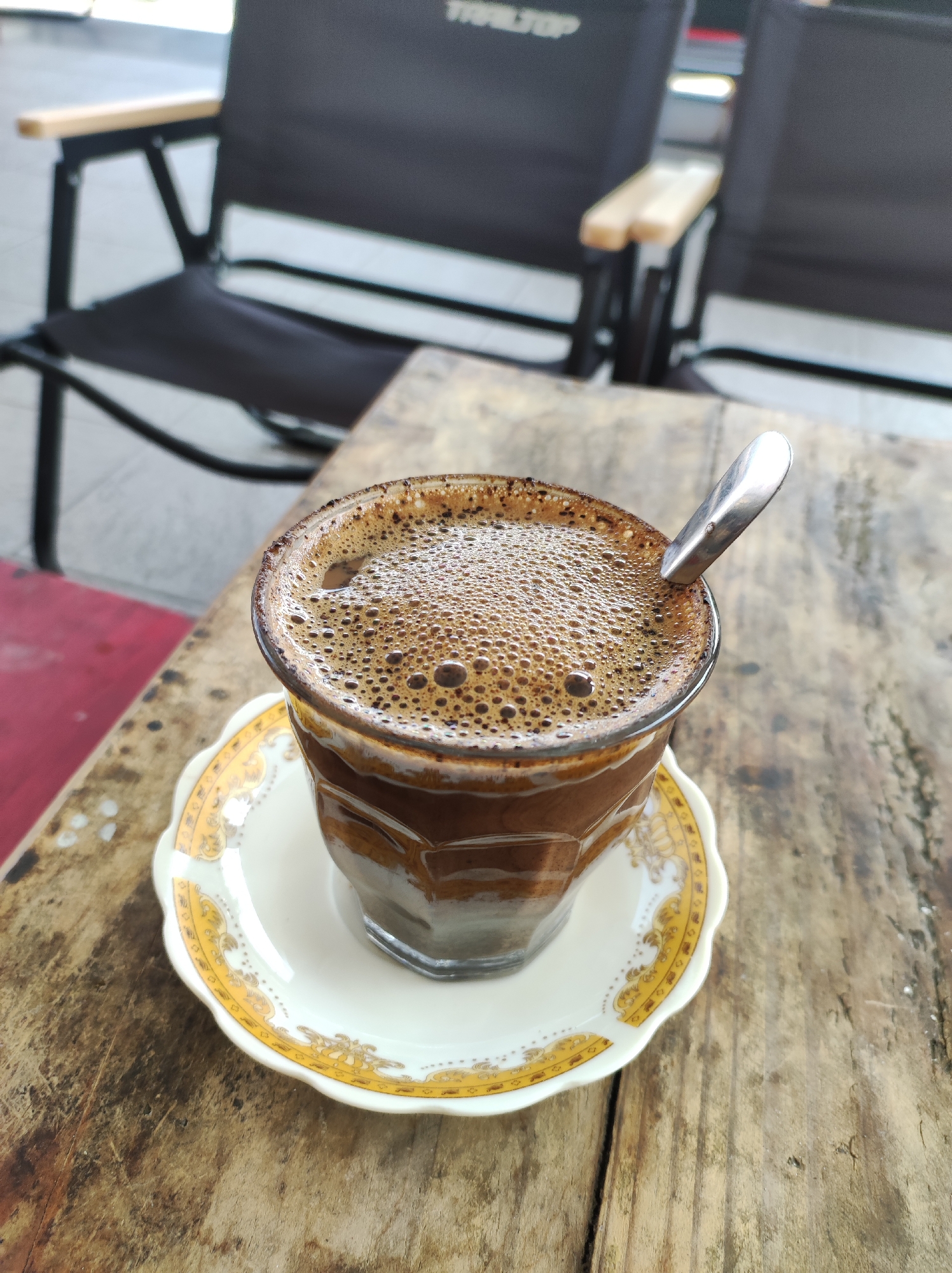 A glass of coffee sits on a saucer on a wooden table. A spoon rests in the coffee. Two black chairs are visible in the background. The image appears to have been taken outdoors in Blitar, Indonesia.
