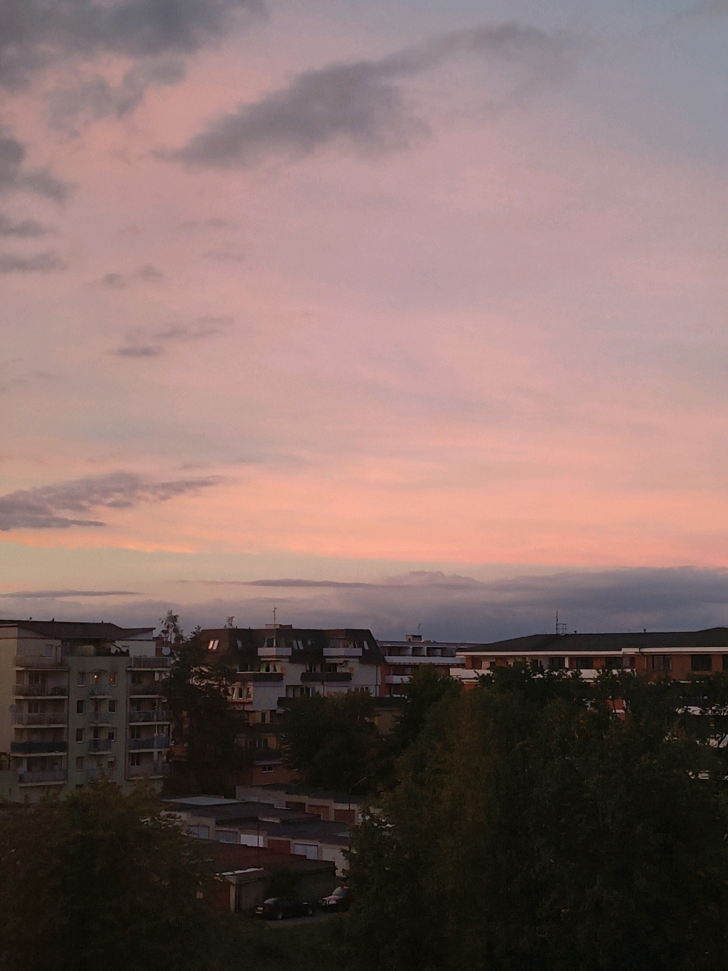 The image captures a dusky sky over a residential area, likely in the city of Olomouc, Czechia. The sky displays a gradient of colors from light pink and orange near the horizon to a muted purple and gray higher up, with scattered clouds contributing to the atmospheric effect. Below the sky, buildings of various heights and architectural styles are visible. A tall, light-colored apartment building with multiple balconies dominates the left side of the frame. To its right, a series of lower-rise buildings with darker roofs and some visible windows are present. Dense foliage of trees partially obscures some of the lower structures. The scene suggests an evening setting, possibly around dusk or twilight, as indicated by the subdued lighting and the coloration of the sky. The overall impression is a quiet urban landscape under a colorful sky.