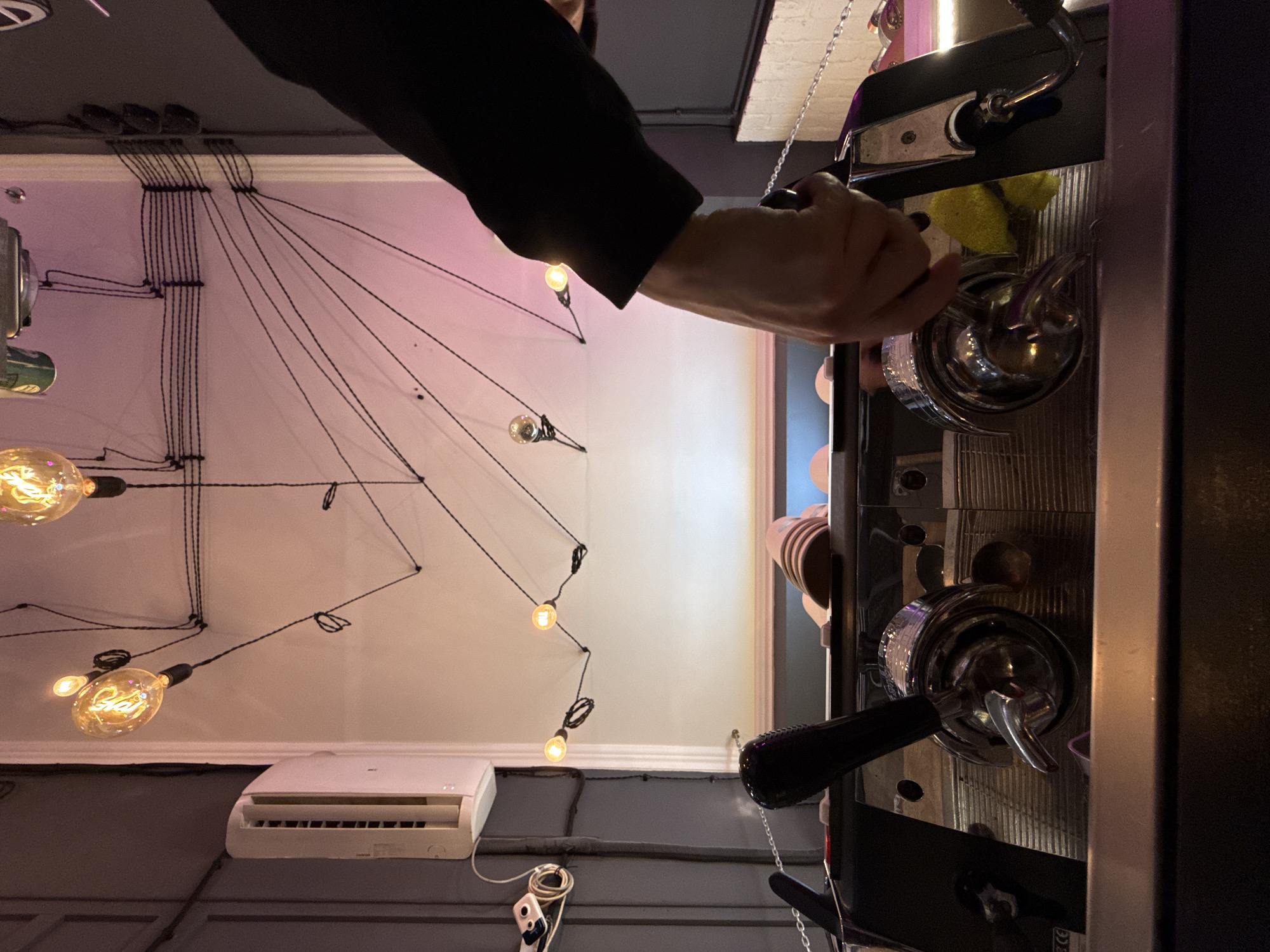 The image depicts an interior scene, possibly a cafe or bar, with a focus on a professional espresso machine. The espresso machine is made of stainless steel and has two portafilters. A person's arm and hand are visible, with the hand appearing to be interacting with the machine, possibly preparing a drink. Above the machine, a network of black wires is strung across a white wall, supporting several Edison-style light bulbs, some of which are illuminated, casting a warm glow. An air conditioning unit is mounted on a gray wall to the left of the frame. The overall ambiance is modern and dimly lit, with a pink hue emanating from some of the lights. There is no information in the image to suggest the location is Moscow, Russia.