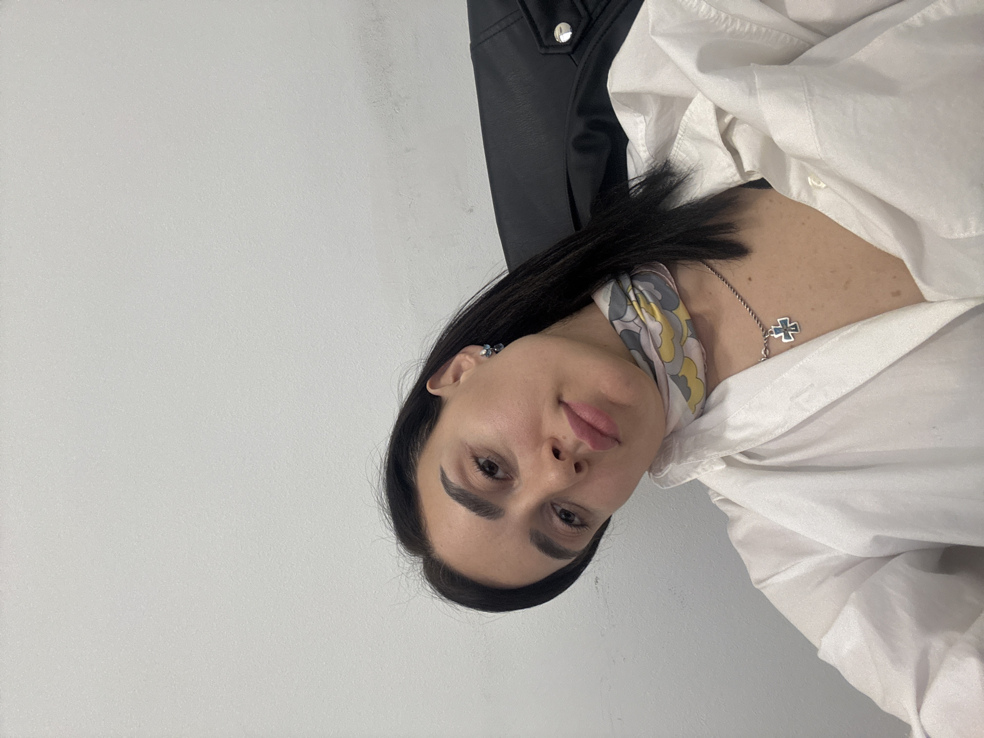 A medium close-up shot shows a woman with dark hair sitting in a chair. She wears a white shirt, a patterned scarf, and a necklace with a cross pendant. A black leather jacket is visible behind her. The background is a plain, light-colored wall. The location is not discernible from the image.