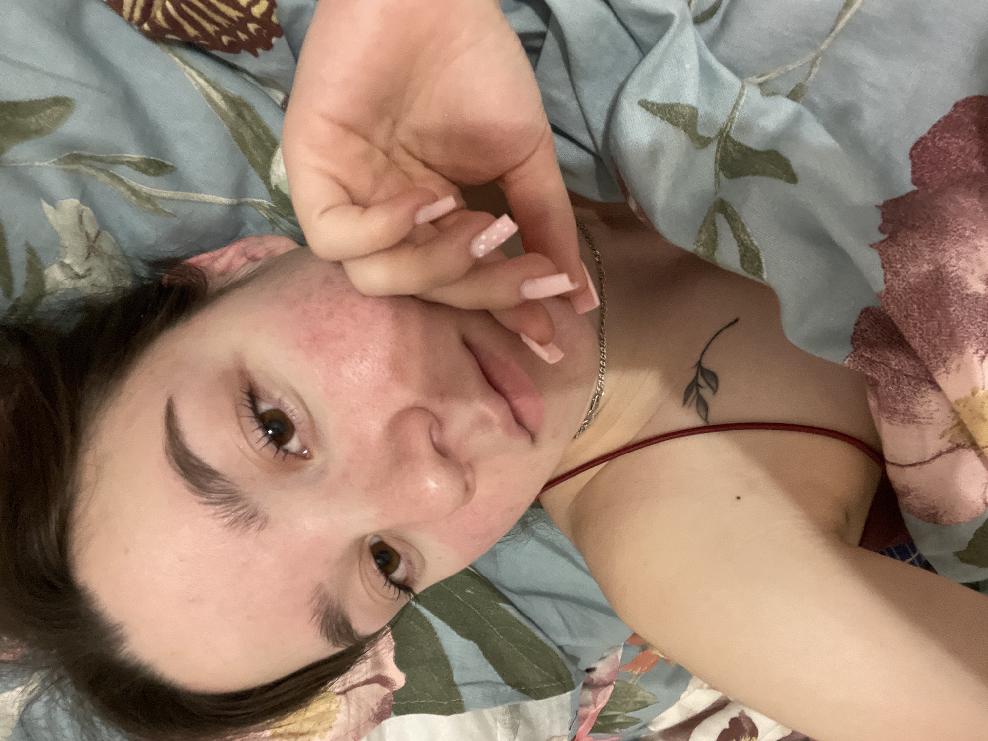 A young, light-skinned woman with brown hair is lying on her back on a floral patterned fabric. She has light pink manicured nails. Her hand is raised near her face. The woman has brown eyes and visible freckles/redness on her face. A silver chain necklace is visible around her neck, along with a small tattoo of leaves on her upper chest. She is wearing a thin-strapped top with a red strap. The background fabric has a blue and beige floral pattern. There is no information available about whether the woman is in Ольховка, Russia.