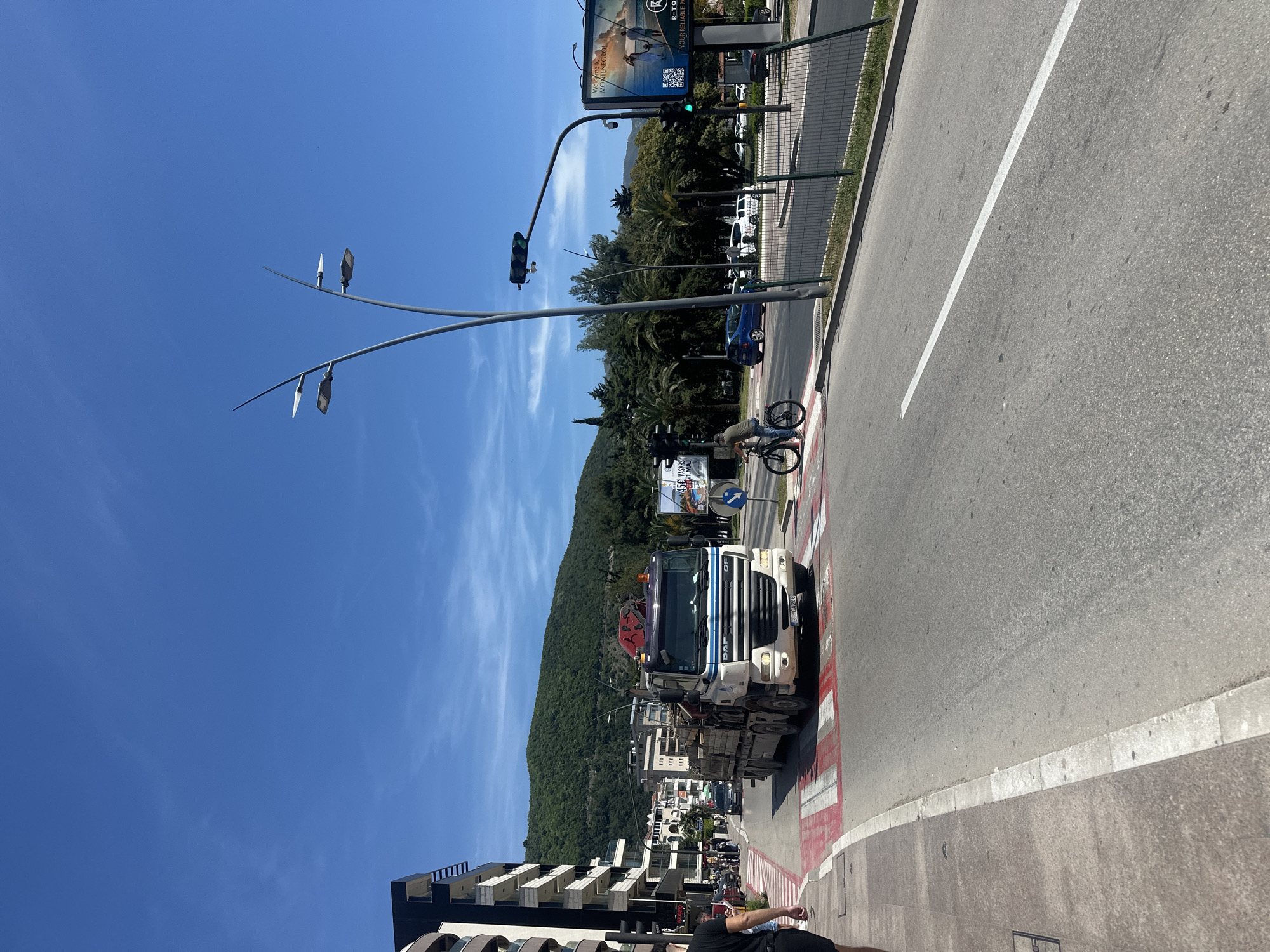The image captures a vibrant daytime street scene in Budva, Montenegro, under a clear blue sky with sparse clouds. The shot is taken from a slightly elevated perspective, looking down a wide urban road flanked by buildings and a lush, tree-covered mountain in the background.
In the foreground, a large white DAF truck, bearing the license plate "BD CA246" and equipped with a crane, is visible on the street, seemingly at an intersection or pedestrian crossing marked with red and white stripes. To the right of the truck, a person on a bicycle is positioned near the sidewalk. The road itself features a clear white dividing line.
Along the right side of the road, a sidewalk and a green mesh fence run parallel to the street. Beyond the fence, several cars, including a blue one, are parked or driving. Prominently featured above the street is a traffic light, currently showing green, and a modern streetlight with multiple lamps. A large billboard advertises "Welcome to MONTENEGRO!" along with text "R-TO" and "YOUR RELIABLE PA," and a QR code. Another sign below it mentions "45€ VASKRS" and "11. MAJ."
Further down the street on the left, modern buildings with multiple stories and balconies are visible. The backdrop is dominated by a majestic, tree-covered mountain range, adding a natural element to the urban setting. The bright sunlight suggests good weather conditions.