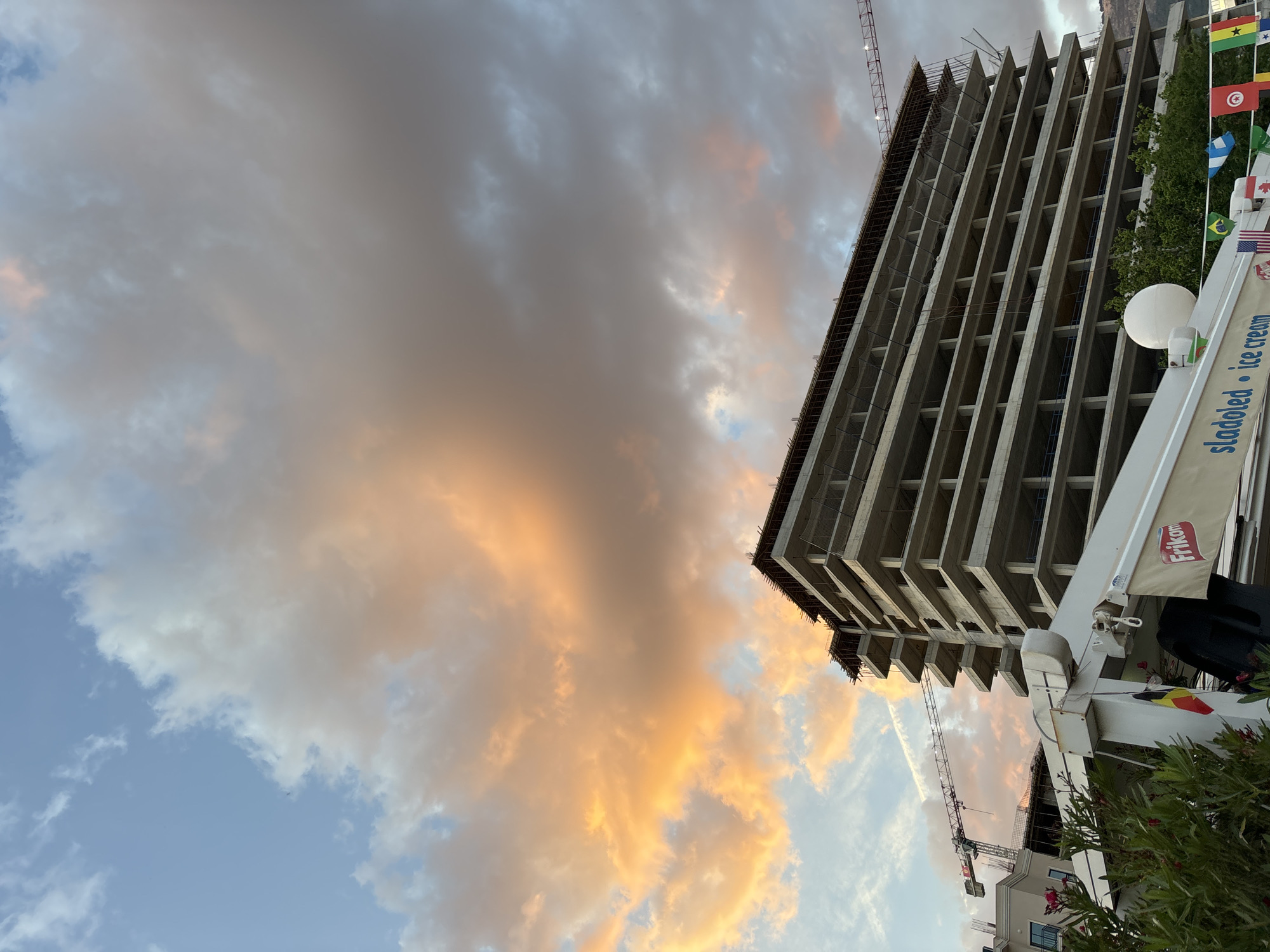 A construction site dominates the scene in Budva, Montenegro. An unfinished high-rise building with multiple open levels stands against a cloudy, sunset-tinged sky. A crane is visible next to the building, suggesting ongoing construction. Flags of various nations are visible attached to a nearby structure. An ice cream stand with "Sladoled Ice Cream" written on its canopy is visible near the base of the frame.