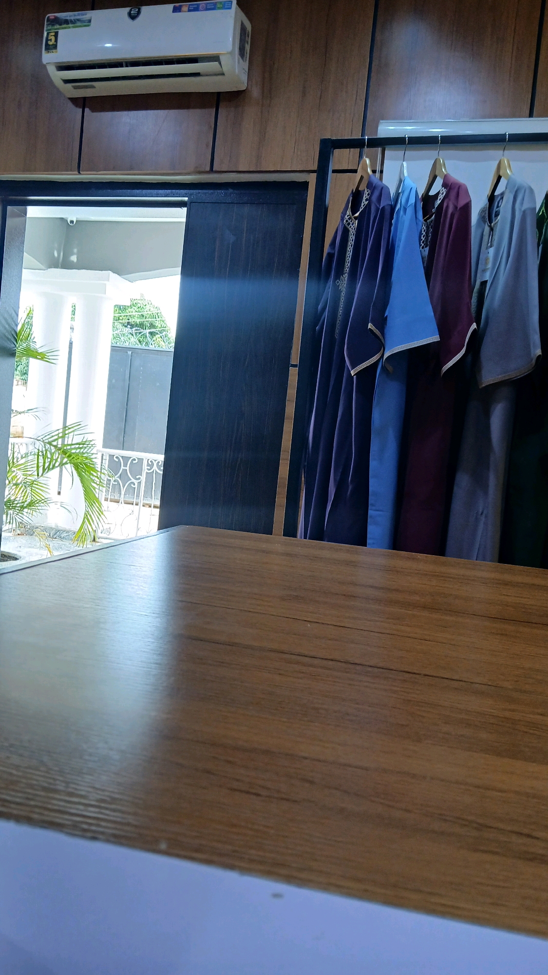 The image shows a close-up view of a wooden table in the foreground, with a rack of traditional Islamic clothing hanging in the background. The clothing consists of several long, flowing robes in various colors, including purple, blue, maroon, and gray. The robes appear to be made of a light fabric and are decorated with ornate embroidery. The background also features an air conditioning unit mounted on a wooden wall, and a partially open doorway leading to an outdoor area with plants and a white railing. The scene is indoors, likely in a retail setting or a home. The location is specified as Gura Top, Nigeria. There are no people visible in the image.