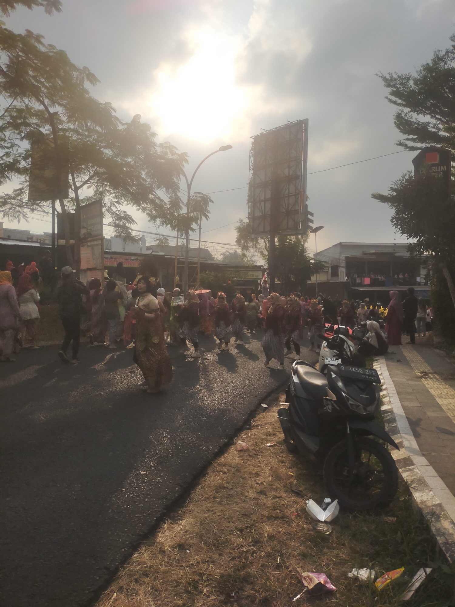 The image depicts a street scene in Tuwel, Indonesia, with a group of people participating in what appears to be a parade or cultural event. The sun is low in the sky, casting long shadows and creating a bright, hazy atmosphere. People are dressed in colorful traditional attire, some wearing head coverings and elaborate costumes. They are walking on a paved road, with a sidewalk bordering the right side of the image. On the sidewalk, a black motorcycle is parked. The environment includes trees, buildings in the background, and a large billboard. There is also some dry grass and scattered litter along the edge of the road and sidewalk. The overall impression is of a community gathering, possibly a celebration or festival.