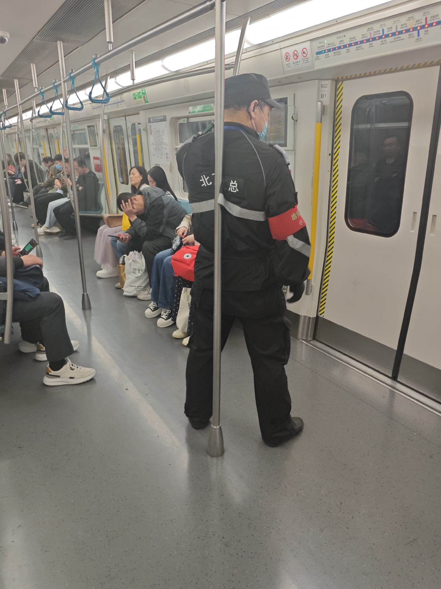 This image captures the interior of a subway train car, bustling with passengers. The central subject is a person in a dark uniform, likely a staff member or security guard, standing facing away from the camera. They wear a black cap, a light blue surgical mask, and a black jacket with grey reflective stripes on the arms and back. On the back of the jacket, Chinese characters "北" and "总" are visible, while a red armband on their left arm displays "治安" (public security) and a small "e" or number "2". They also have a small red bag strapped to their side.
Numerous other passengers are visible, mostly seated along the sides of the car. Some are engrossed in their smartphones, while others appear to be resting or observing their surroundings. One passenger is notably seen with their head in their hands, suggesting tiredness or distress. The subway car features a grey floor, white walls, overhead handles with blue straps, and bright overhead lighting. A yellow vertical pole with black diagonal stripes stands beside a closed train door, which is marked "M2 B1". Above the windows, various emergency signs and a route map with station names are displayed. Partially visible Chinese text on the map includes "门" and "北京站" (Beijing Station), suggesting the location is Beijing, China. A white tote bag belonging to a passenger clearly shows the text "金所" and "81776687". The overall atmosphere is that of typical urban commuting.