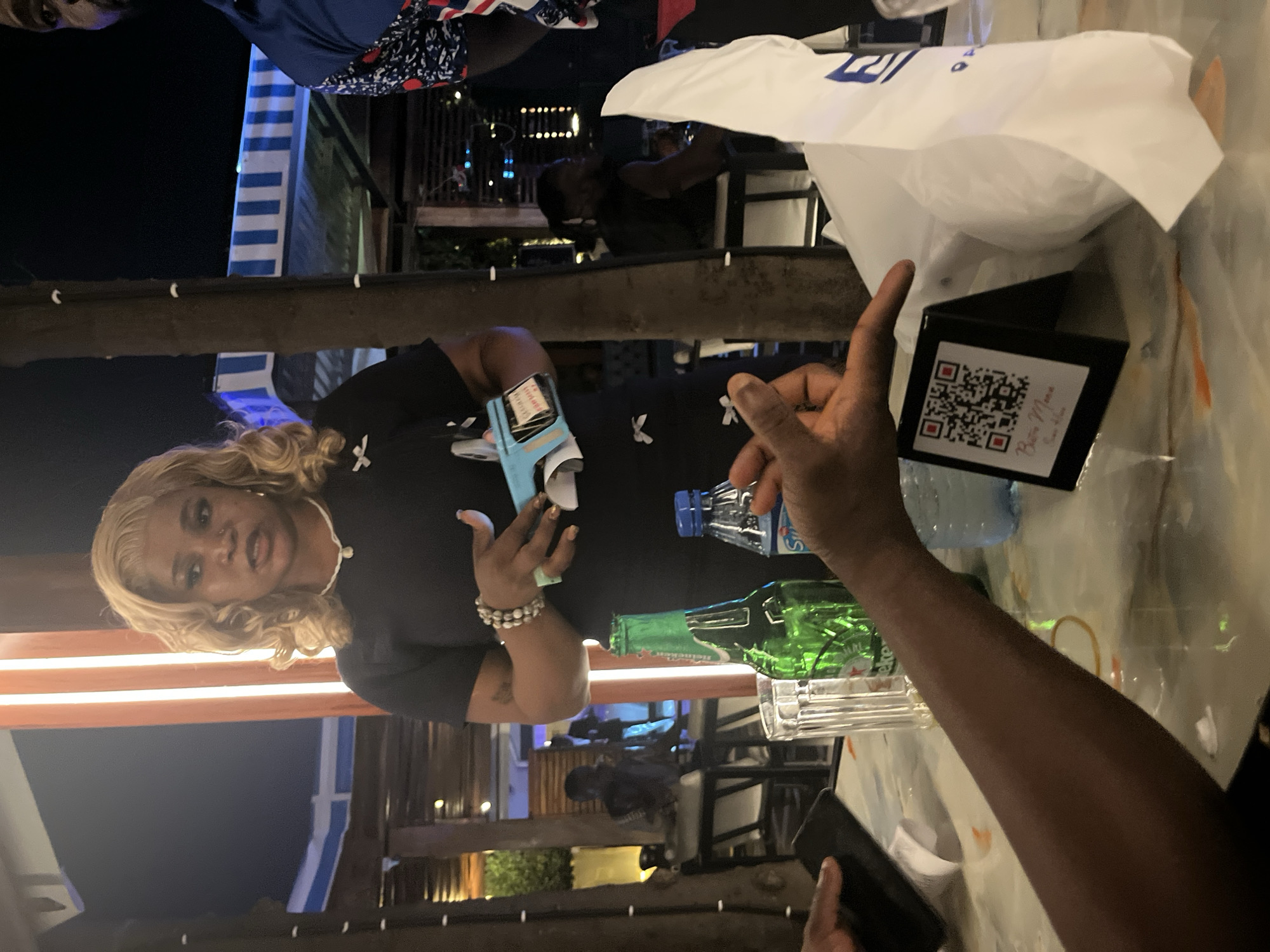 This image depicts a nighttime scene at an outdoor seating area, possibly a restaurant or bar, in Abuja, Nigeria. A woman with blonde, curly hair, wearing a dark dress adorned with white bow-like patterns, is the central figure. She is holding a light blue object and is in the process of speaking or interacting with someone off-camera. In the foreground, a table is visible with several items, including a green glass bottle (likely a beverage), a glass of water, and a plastic bottle of water. A hand, likely belonging to another person, is reaching towards the table. To the right, a white paper bag and a black stand with a QR code are prominent. The background shows additional seating areas, trees, and lighting, suggesting a lively atmosphere. The presence of the QR code and menu item name suggests the location is a dining establishment. The overall impression is a casual gathering or dining experience.