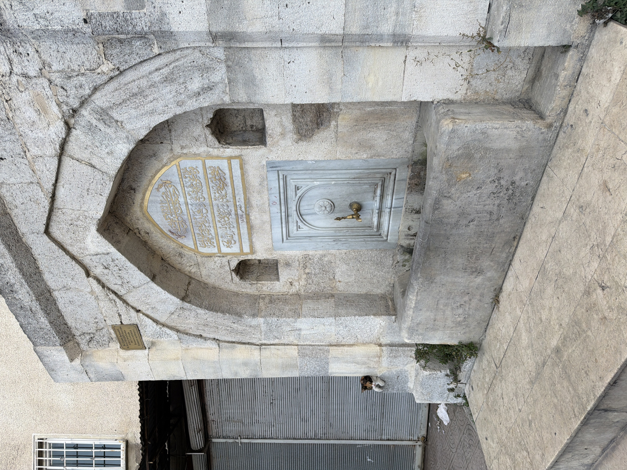 The image depicts an outdoor public fountain in Istanbul, Turkey. The fountain is constructed of aged stone and features an arched alcove with decorative Islamic calligraphy above a marble panel containing a brass faucet and a carved floral motif. Below the faucet is a trough for collecting water. To the left of the fountain is a closed metal shuttered shopfront, and to the right, part of a building with a barred window is visible. A small, calico cat is perched on a ledge to the left of the fountain, near the shuttered storefront. The ground in front of the fountain is paved with stone tiles.