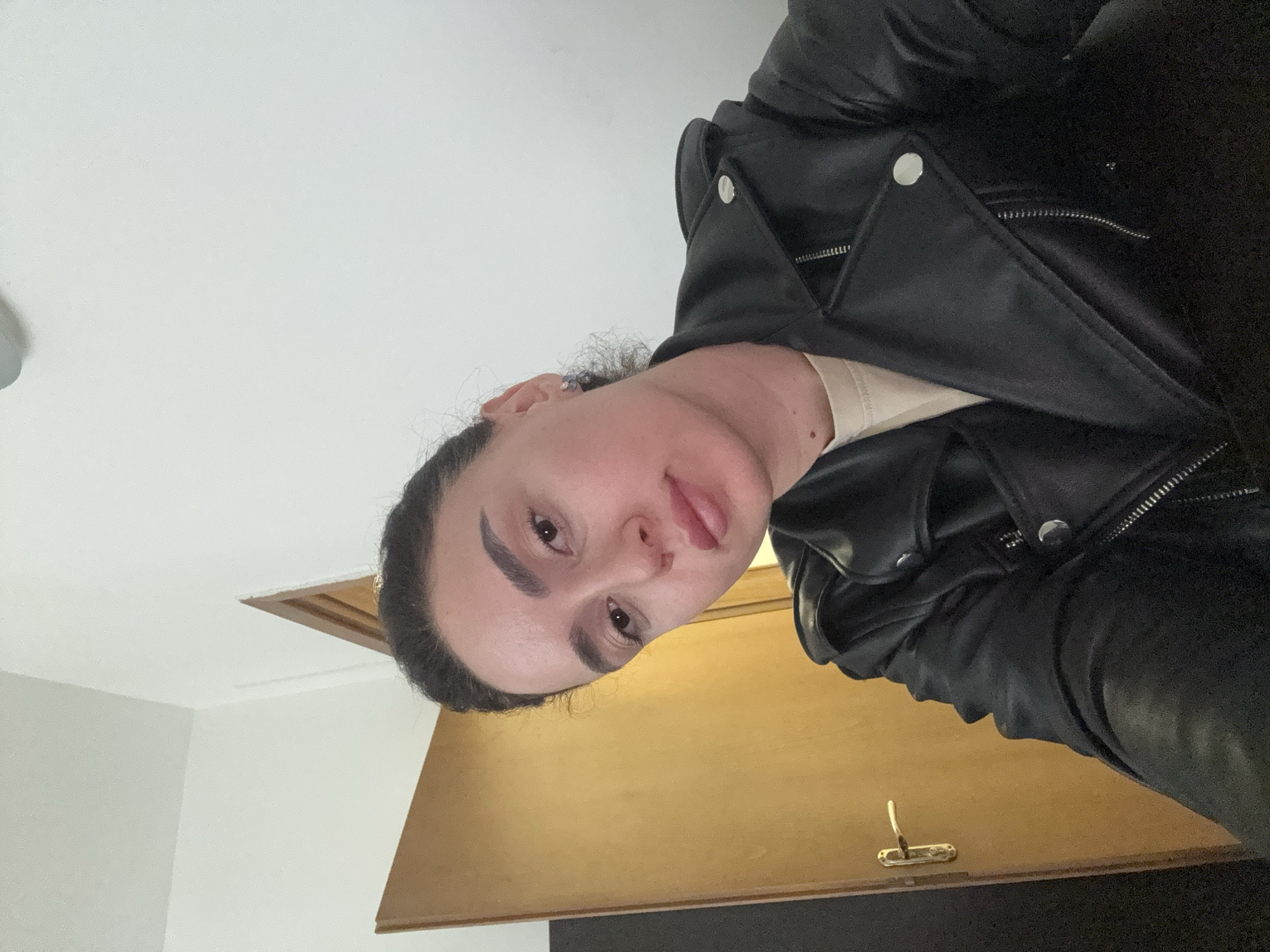 A person wearing a black leather jacket is in a room with white walls and a wooden door. The door has a brass handle. The person has dark hair pulled back and is sticking out their tongue. The location cannot be determined without additional information.