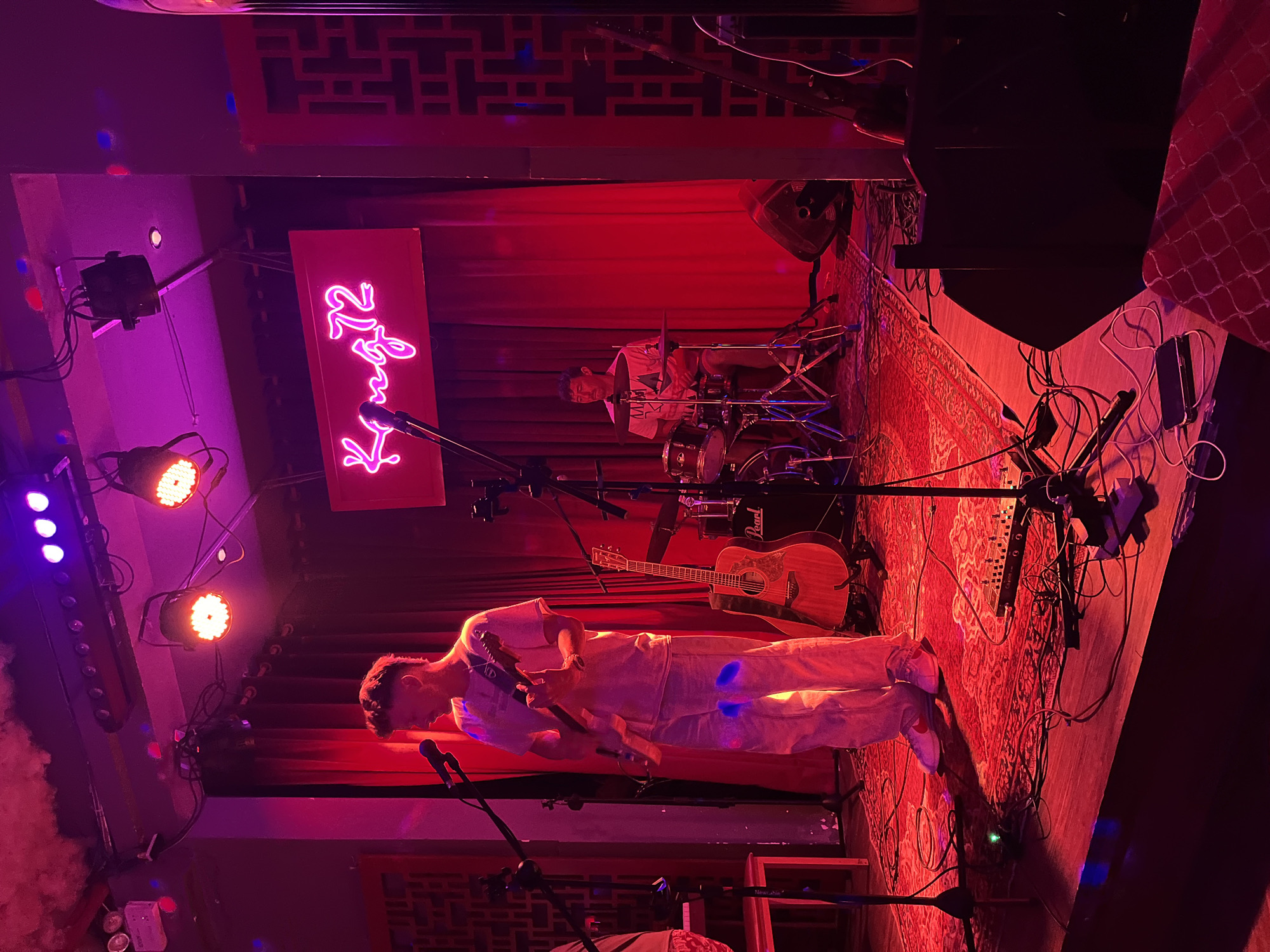 The scene depicts a band performing on a small stage in a dimly lit venue with red and purple lighting. A male guitarist, wearing a white outfit, is in the foreground, playing an electric guitar. Behind him, a drummer is seated at a Pearl drum set. An acoustic guitar is resting on the floor. The stage is covered with a red patterned rug. Behind the drummer, there is a red curtain. A neon sign is visible, possibly reading "Kyo 72." The lighting equipment is visible on the sides of the stage. Wires and cables are strewn across the stage floor. The venue appears to be in Đà Nẵng, Vietnam, as indicated by the name "Kyo 72" which is the location's name.