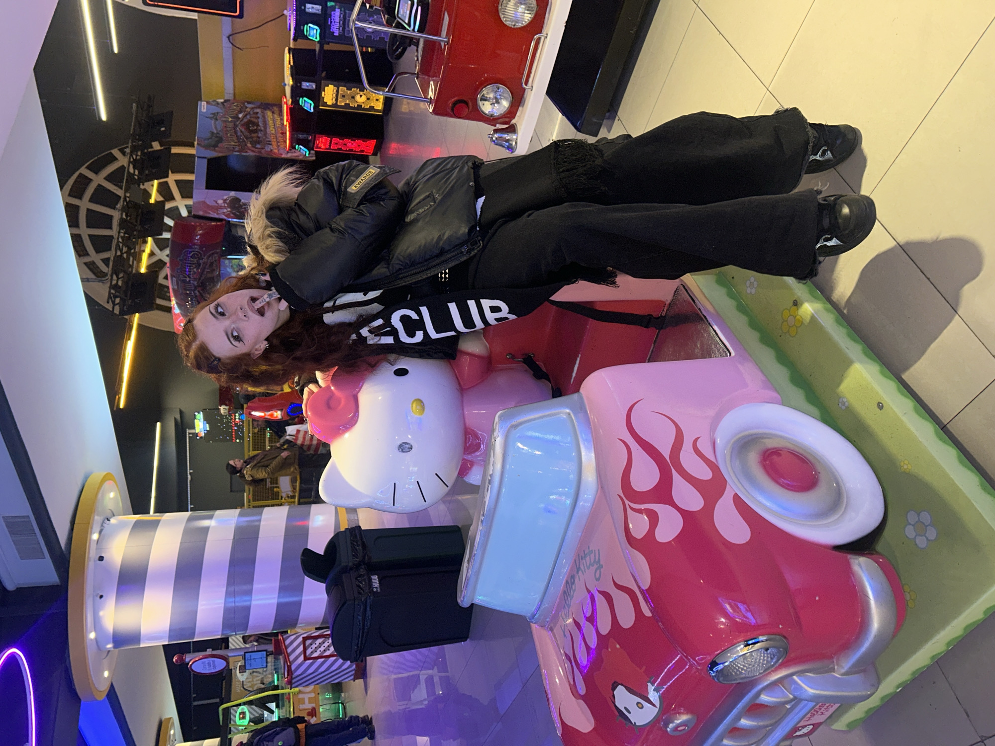 A young woman with long, auburn hair and a white and black scarf around her neck sits on a pink Hello Kitty themed amusement park ride. She is wearing a black puffer jacket with a fur-lined hood and wide-legged black pants. The ride is shaped like a retro car with flames. In the background, there are several arcade games with colorful lights. The scene appears to be set in an indoor amusement center or arcade, possibly in Buenos Aires, Argentina, given the context. The lighting is dim and colorful, typical of such venues.