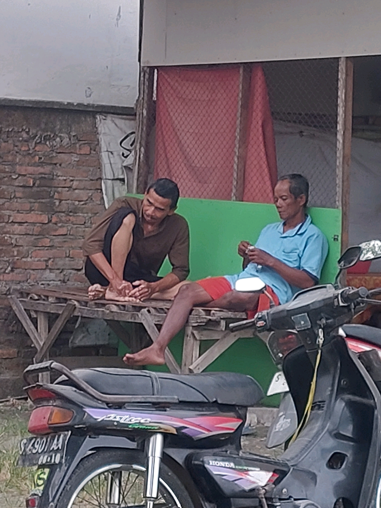 Two adult males are seated on a wooden bench in an outdoor setting, likely in Sarangan, Indonesia. The man on the left is wearing a brown long-sleeved shirt and dark pants, and is crouched with his legs bent and one foot resting on the bench. He appears to be examining his feet. The man on the right is wearing a light blue short-sleeved collared shirt and red shorts. He is holding a small white object in his hands, possibly paper, and looking down at it. Both men have short, dark hair and appear to be of Indonesian descent. Behind them is a green structure with a red mesh covering. In the foreground, a black motorcycle is parked. The background shows a brick wall on the left and hints of a building structure. The ground appears to be dirt or gravel.