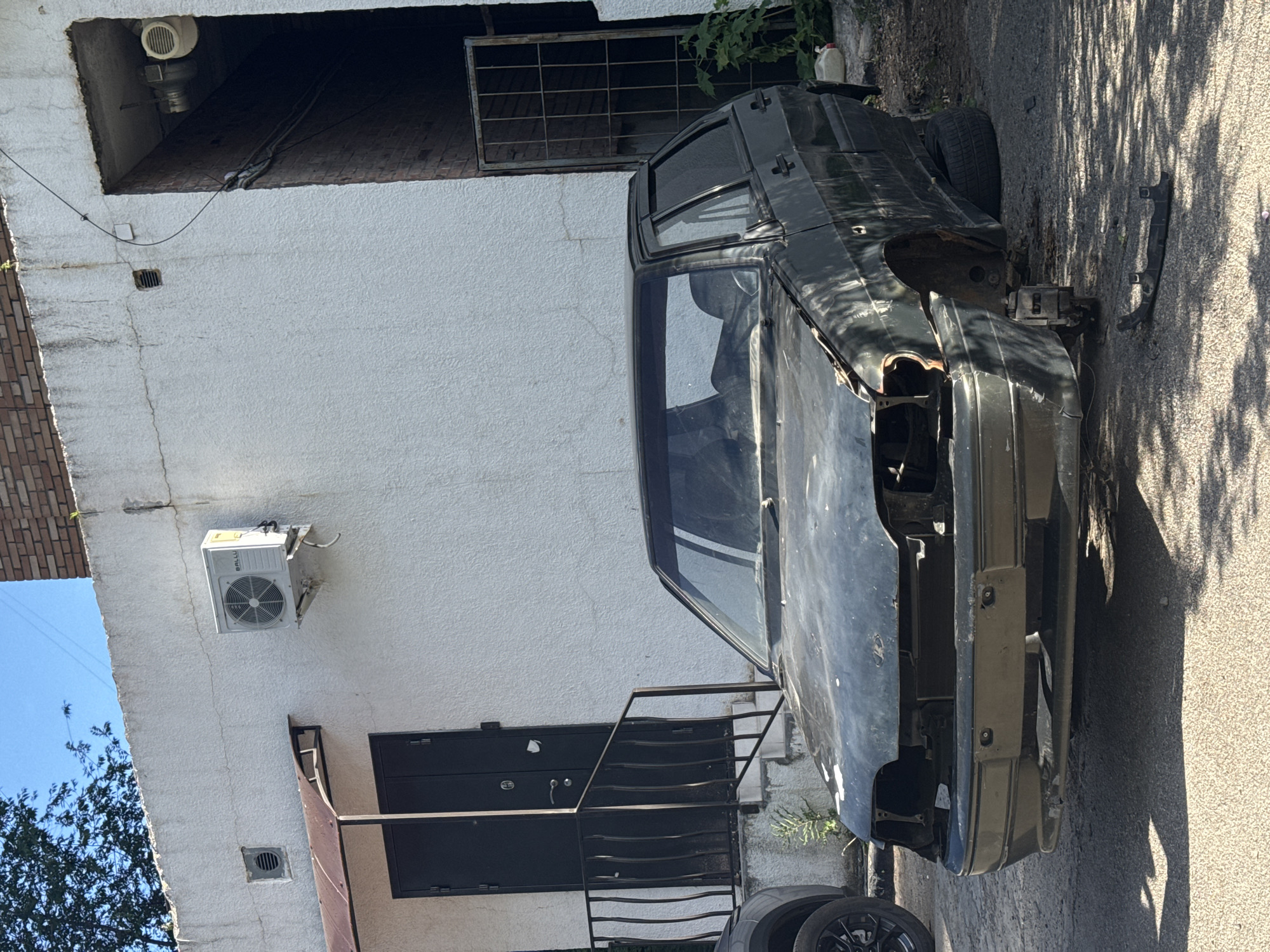 A high-angle, outdoor shot captures a heavily damaged, dark-colored car parked alongside a white, textured building under bright daylight. The car, possibly a sedan or hatchback, is extensively stripped, missing its front bumper, headlights, and other components, revealing rust and exposed mechanics. Its body panels show wear and paint peeling, and the windows reflect the sky. A single tire is visible at the rear of the vehicle, with another loose wheel and various car parts scattered on the paved ground nearby.
The white building features a gridded window and a dark double door accessible via a small metal railing or staircase. Mounted on the wall above the door is a white air conditioning unit, clearly displaying the brand name "BALLU". Faint cracks are visible on the building's textured white facade. To the left, a section of an older brick building is seen, partially obscured by tree branches. The ground is paved with asphalt or concrete, with harsh, dappled shadows from unseen trees and the car itself, indicating a sunny day. The scene evokes a sense of abandonment or a vehicle awaiting salvage in an urban or industrial setting. No people are present.