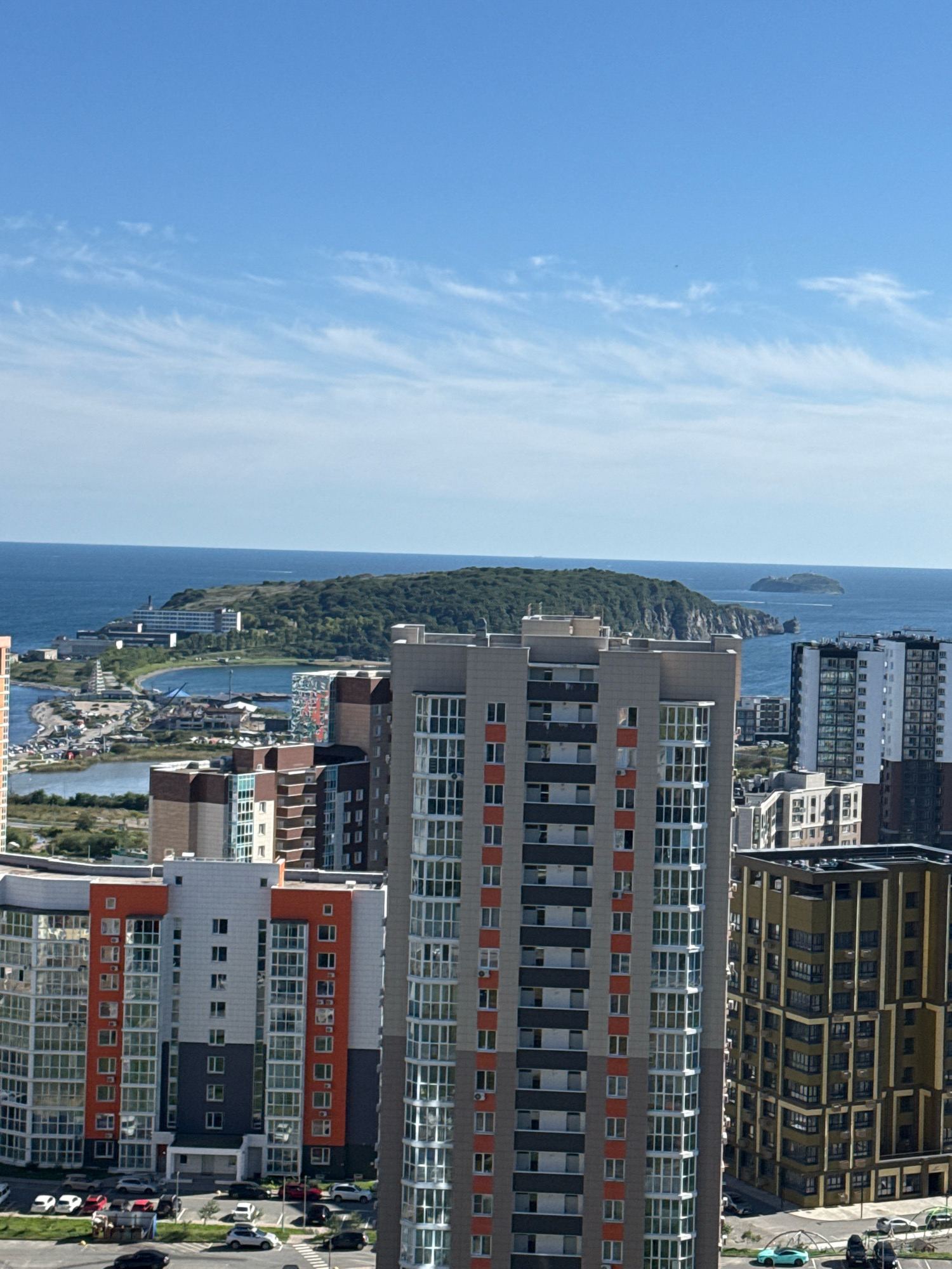 The image is an aerial view of a coastal city with several high-rise buildings in the foreground. In the background, there is a large body of water with a green, tree-covered island in the middle distance. The sky is blue with some scattered white clouds. The buildings in the foreground are modern apartment complexes with varying architectural styles and colors, including orange, grey, and white facades. Some buildings have many windows, indicating residential units. Below the buildings, a parking lot with several cars is visible, along with roads and some greenery. The scene suggests a developed urban area adjacent to a natural coastal landscape. The buildings are consistent with the modern architecture found in Vladivostok, Russia, particularly in areas with new residential developments.