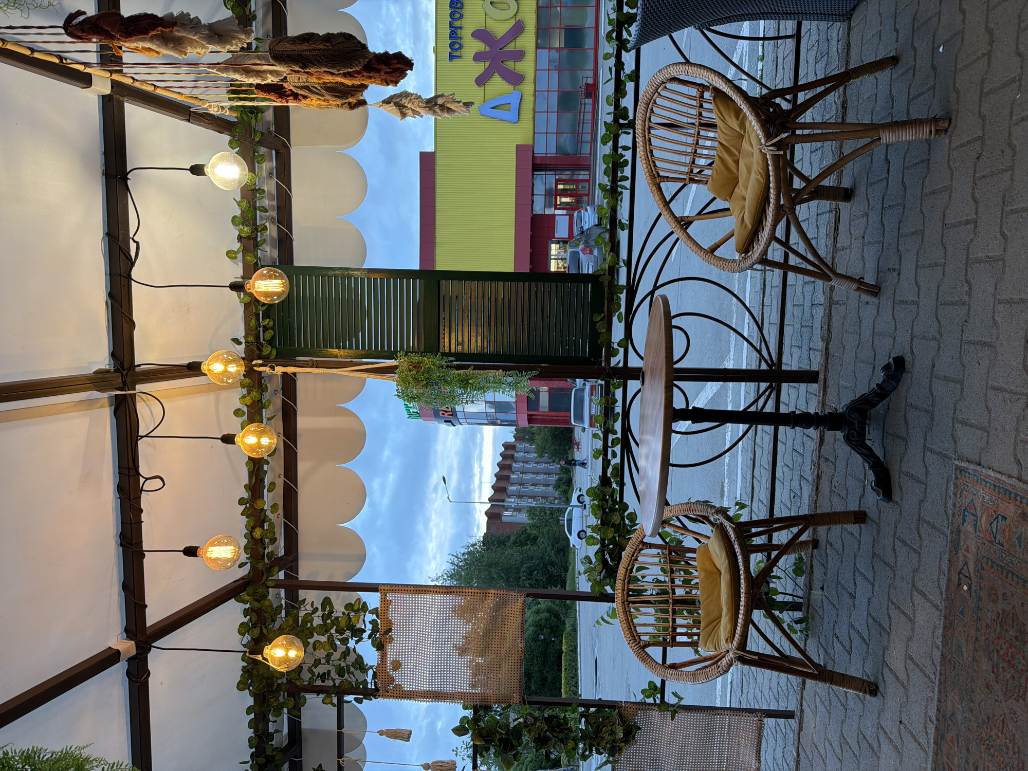 This image captures an outdoor, covered seating area of a cafe or restaurant in Chaikovsky, Russia, at dusk. The scene looks out onto a street and several buildings, most notably the "ДЖОНКА" (Jonka) shopping center.
In the foreground, two light-brown wicker chairs, each fitted with a mustard-yellow cushion, are positioned around a small, circular wooden table with a black, ornate metal base. The floor is paved with grey interlocking pavers, partially covered by a rectangular, earth-toned patterned rug. A decorative black metal railing with scrollwork separates the seating area from the street. Overhead, a white fabric awning with scalloped edges provides cover, supported by dark brown structural beams. Numerous antique-style, spherical filament light bulbs hang from black cords, emitting a warm glow. Artificial green leafy vines are draped along the structural beams. To the right, various hanging decorative elements made of textured fibers or macrame in shades of brown, beige, and orange are suspended from a wooden beam. A dark green louvered panel and a woven rattan panel are visible as vertical partitions.
In the midground, beyond the railing, is an asphalt road or parking lot. A white sedan is parked towards the left, and a darker sedan is partially visible on the right. A single individual is discernible walking in the distance on the far side of the road, near a lamppost.
The background is dominated by a large commercial building painted bright yellow with red trim around its windows and entrance. A prominent blue sign on its facade displays the Cyrillic text "ТОРГОВЫЙ ЦЕНТР" (Shopping Center) above "ДЖОНКА". To the left of this building, a multi-story brownish-beige building, possibly residential or office, is partially obscured by trees. The sky above is overcast with a muted blue-grey tone, consistent with twilight conditions.