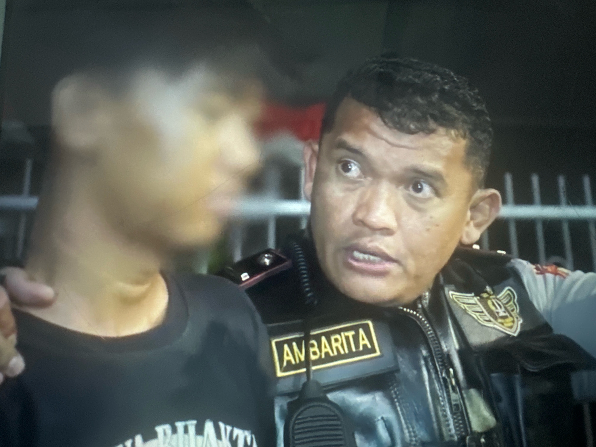 The image shows a close-up of two individuals in what appears to be an outdoor, nighttime setting in the City of Binjai, Indonesia. On the right is a man wearing a dark uniform vest with a name tag reading "AMBARITA" and a police or security agency emblem on the left side of the vest. He has dark hair and is looking to his left with a concerned expression. His mouth is slightly open, as if speaking. His left hand is visible, resting on the shoulder or arm of the person to his left.
The person on the left is blurred and facing away from the camera, wearing a dark t-shirt. Their head and upper body are visible. The background is dark with horizontal bars, possibly a fence, and some blurred lights or objects that are not clearly discernible. The overall impression is of an interaction between a law enforcement or security officer and another person.
