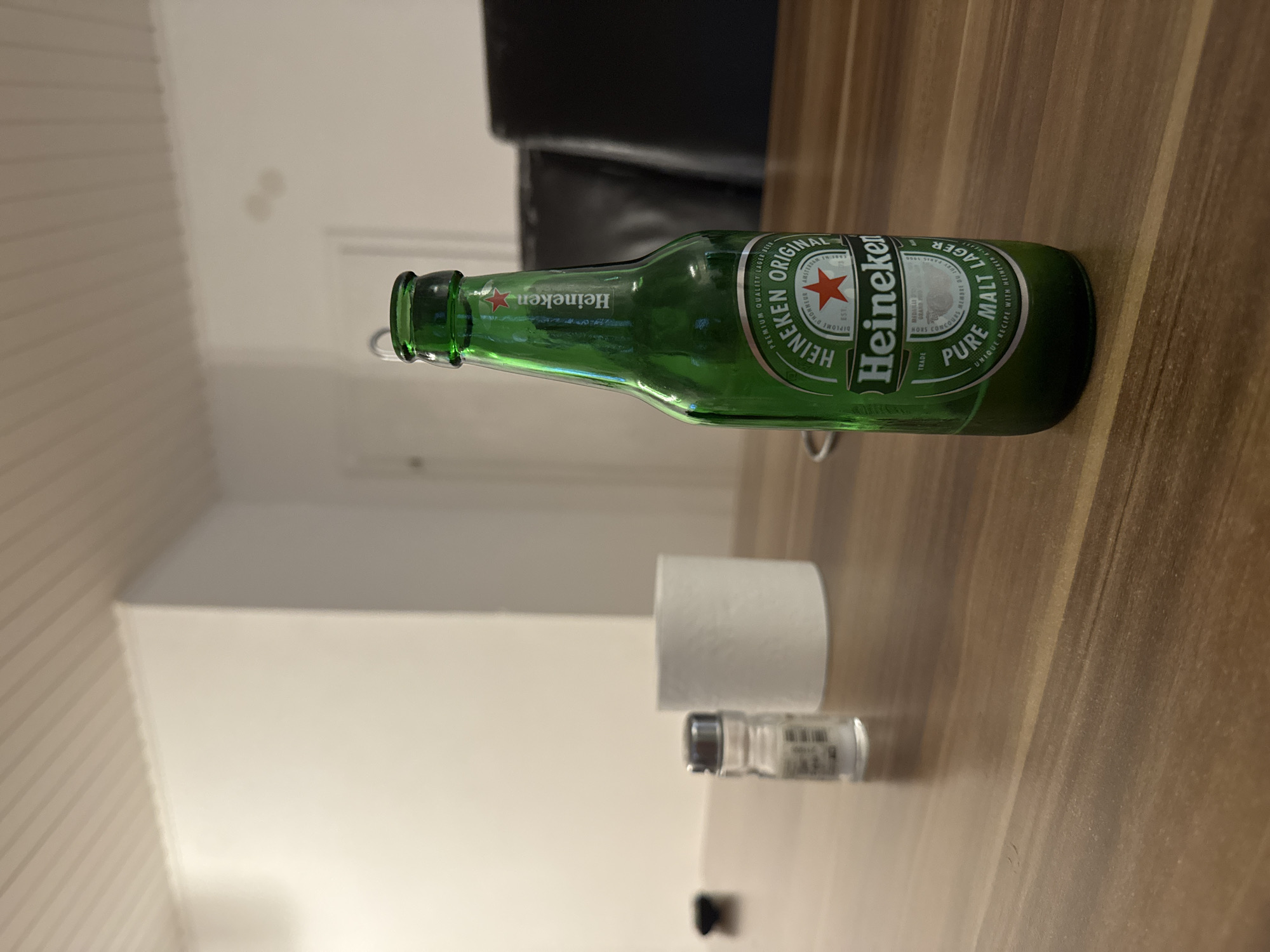 A green Heineken beer bottle lies horizontally on a brown wooden surface, along with a roll of white toilet paper and a small, transparent vial. The image is taken indoors, showing a portion of a chair with black leather, and a room with light-colored walls. The location is Hattersheim am Main, Germany.