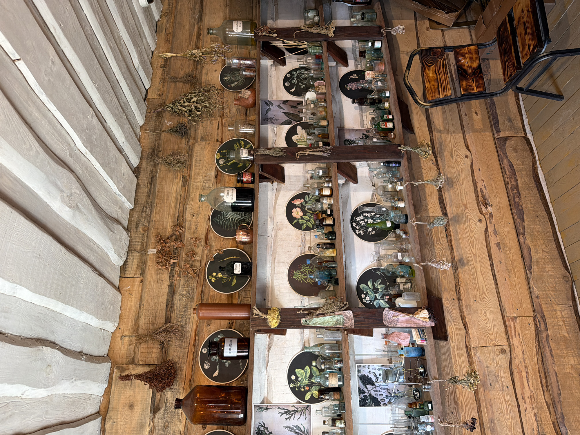 The image shows an interior scene of what appears to be an apothecary or a historical display room, likely located in the Літвенскі сельскі Савет region of Belarus. The walls are constructed from rough-hewn logs, and the ceiling features exposed wooden beams.
The primary focus is on several wooden shelves laden with a collection of antique glass bottles of various shapes, sizes, and colors, some containing liquids. Among the bottles are also decorative plates with botanical illustrations, dried herbs hanging from the walls and shelves, and other assorted objects suggestive of historical medicinal or alchemical practices.
A single, industrial-style metal chair with wooden slats is positioned in the lower right of the frame, facing the shelves. The lighting suggests an indoor environment, possibly with natural light filtering in, creating a rustic and somewhat dimly lit atmosphere. The overall impression is one of preservation of historical artifacts and a specific aesthetic.