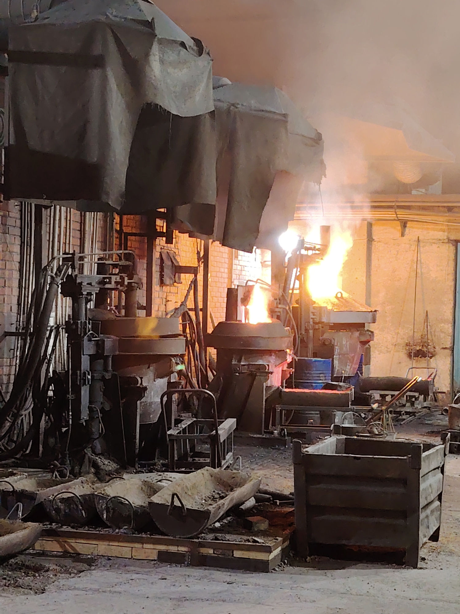 The image shows an industrial setting, likely a foundry or metalworking facility, in Noviy, Russia. There are several pieces of heavy machinery, including furnaces or melting pots emitting flames and smoke, which suggests the presence of molten metal. One area is covered by heavy, draped material. Various containers and troughs are visible in the foreground, potentially used for holding materials. The interior shows exposed brickwork and pipes, indicative of an older industrial building. There are no people depicted in the image. The overall scene conveys a hot, potentially hazardous environment related to high-temperature metal processing.