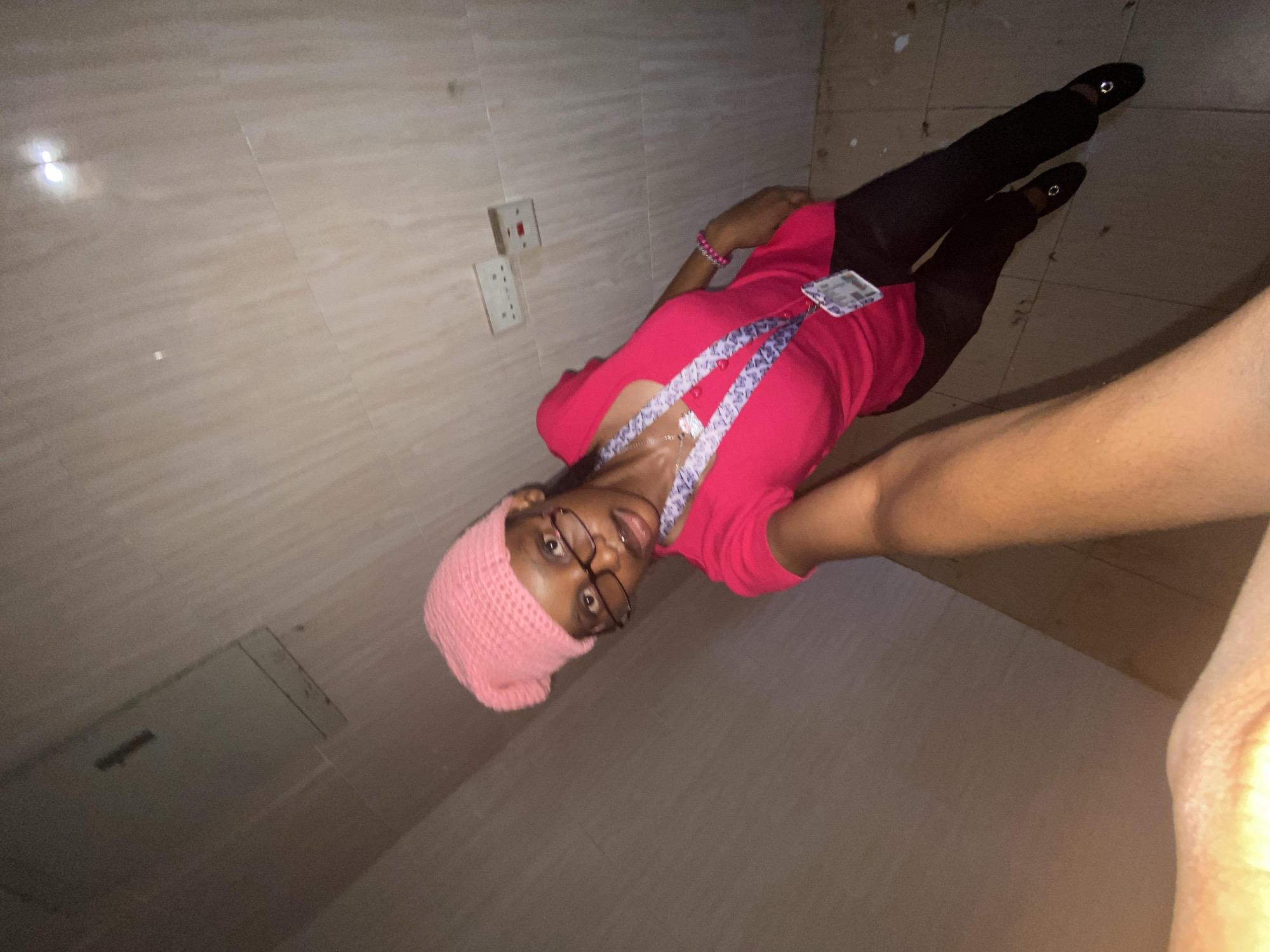 A woman is pictured in a bathroom. She wears a pink beanie, glasses, and a pink shirt. She has on black pants and black shoes. A name badge hangs from her neck. The bathroom has light-colored tiled walls and floor, with electrical outlets on one wall.