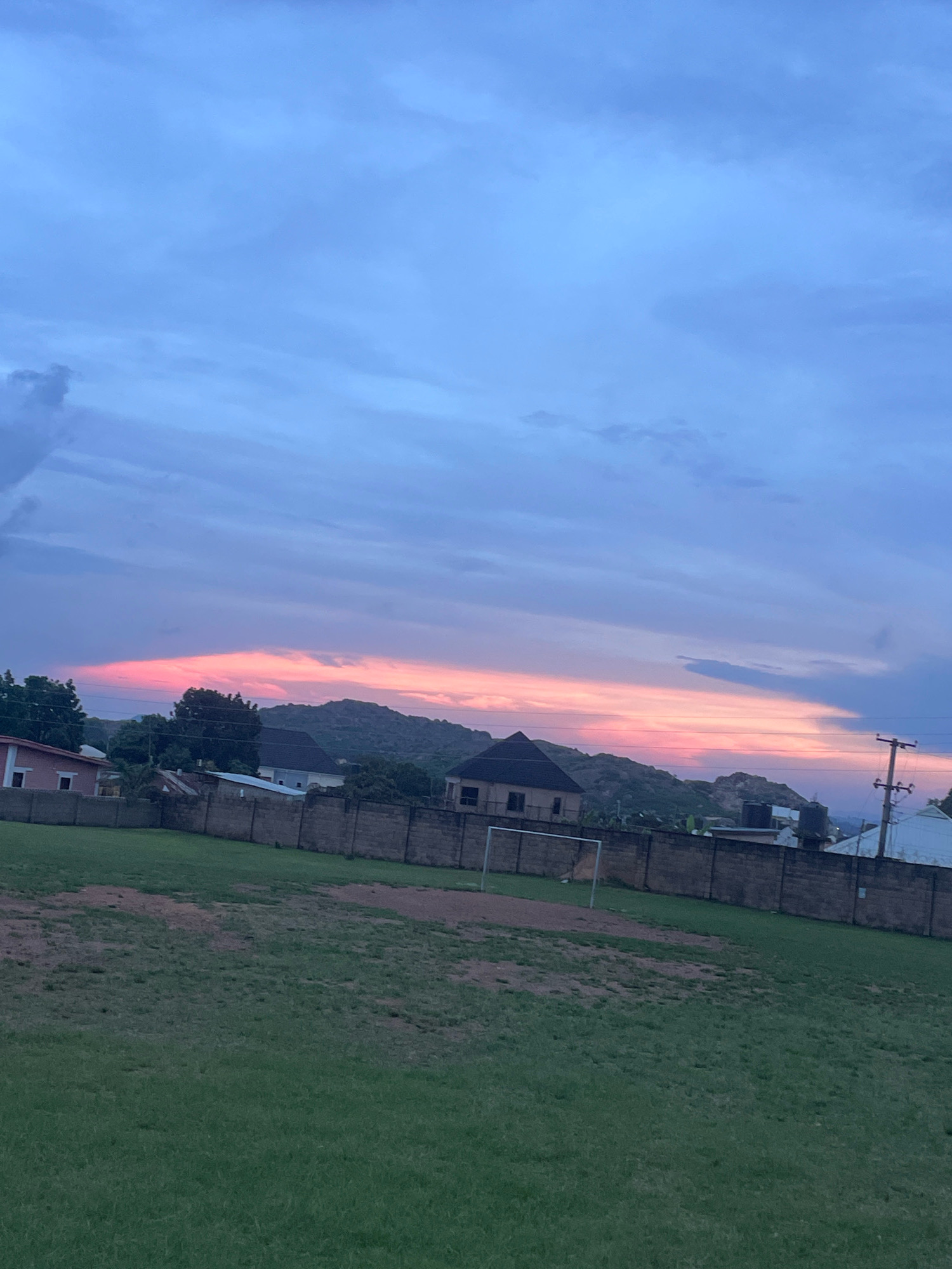 The image depicts a serene outdoor scene at dusk, likely in Jos, Nigeria. The sky is a canvas of soft blues and purples, with a striking band of pink and orange clouds stretched across the horizon, hinting at the setting sun. In the foreground, a well-maintained green field, possibly a sports pitch, occupies the lower half of the frame. A metal soccer goal is visible on the field, suggesting it's a place for recreation.
Beyond the field, a sturdy, light-colored brick wall runs horizontally, serving as a boundary. Behind the wall, several buildings of varying architectural styles can be seen, including a pink and white house on the left, and more modern structures with dark roofs further back. Some of these buildings are nestled against the backdrop of rolling, rocky hills, which are characteristic of the Jos Plateau. A prominent feature on the right side of the frame is a utility pole with electrical wires, indicating the presence of infrastructure. A black water tank is also visible atop one of the buildings. The overall atmosphere is one of tranquility and the quiet beauty of twilight in an urban or semi-urban setting.