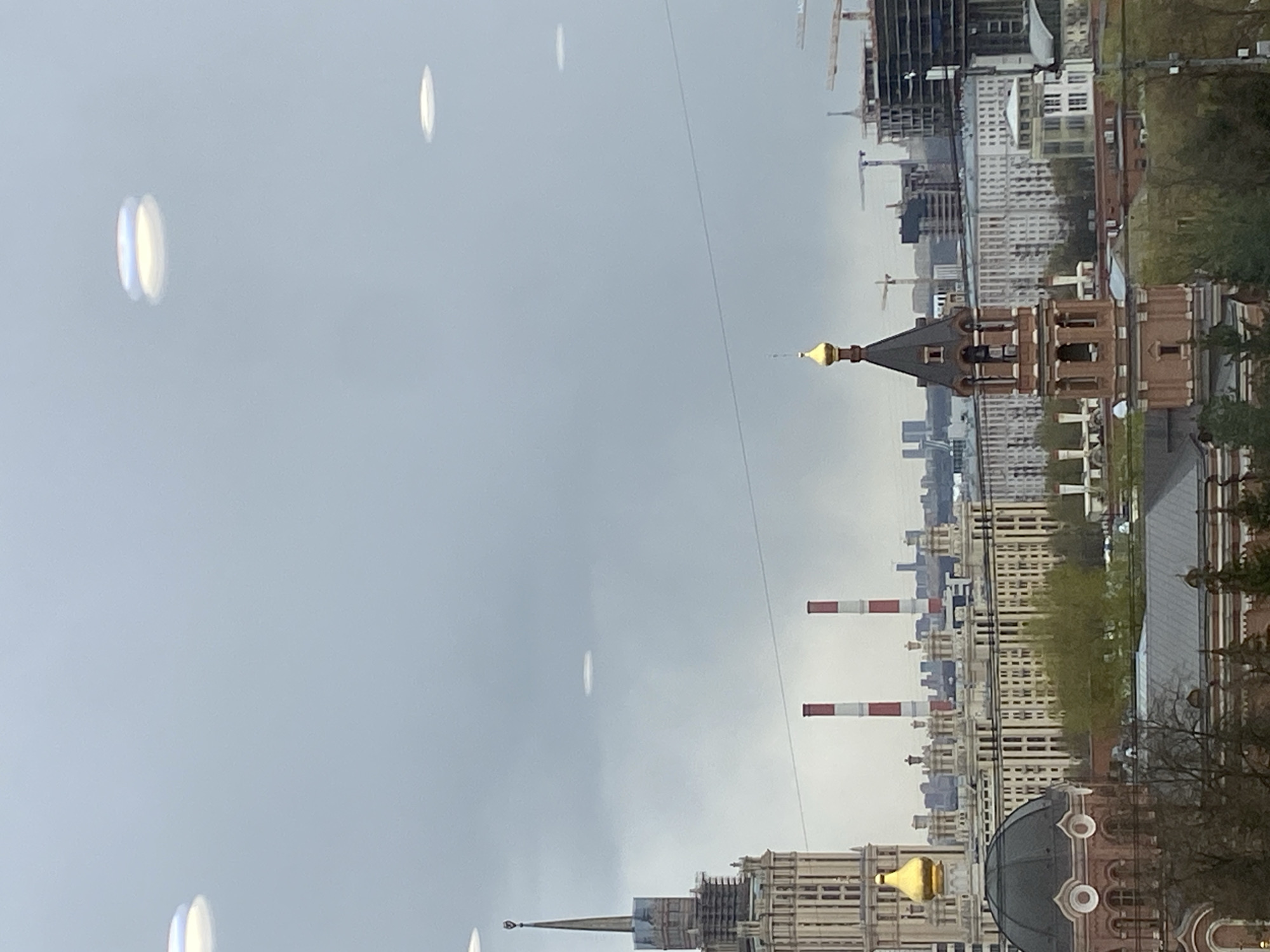 The image displays a panoramic view of a Moscow cityscape under an overcast sky. Dominating the scene is a mix of architectural styles, including several buildings with golden domes and spires characteristic of Russian Orthodox churches. One prominent structure features a golden spire atop a reddish-brown brick building. Another smaller golden dome is visible towards the bottom right of the frame.
The urban landscape includes numerous modern high-rise buildings, with some construction cranes indicating ongoing development. In the mid-ground, two distinct industrial chimneys, striped red and white, rise above the other structures. Patches of trees, some appearing bare and others with green foliage, are scattered throughout the city, suggesting a season other than peak summer or winter.
The sky is uniformly hazy and light grey, indicating an overcast or cloudy day. Several elongated, out-of-focus bright spots are present in the sky, appearing to be lens flares or reflections. A thin, dark line, possibly a wire, stretches diagonally across the sky. The image captures a static, daytime view of the city, devoid of specific human activity or visible text.