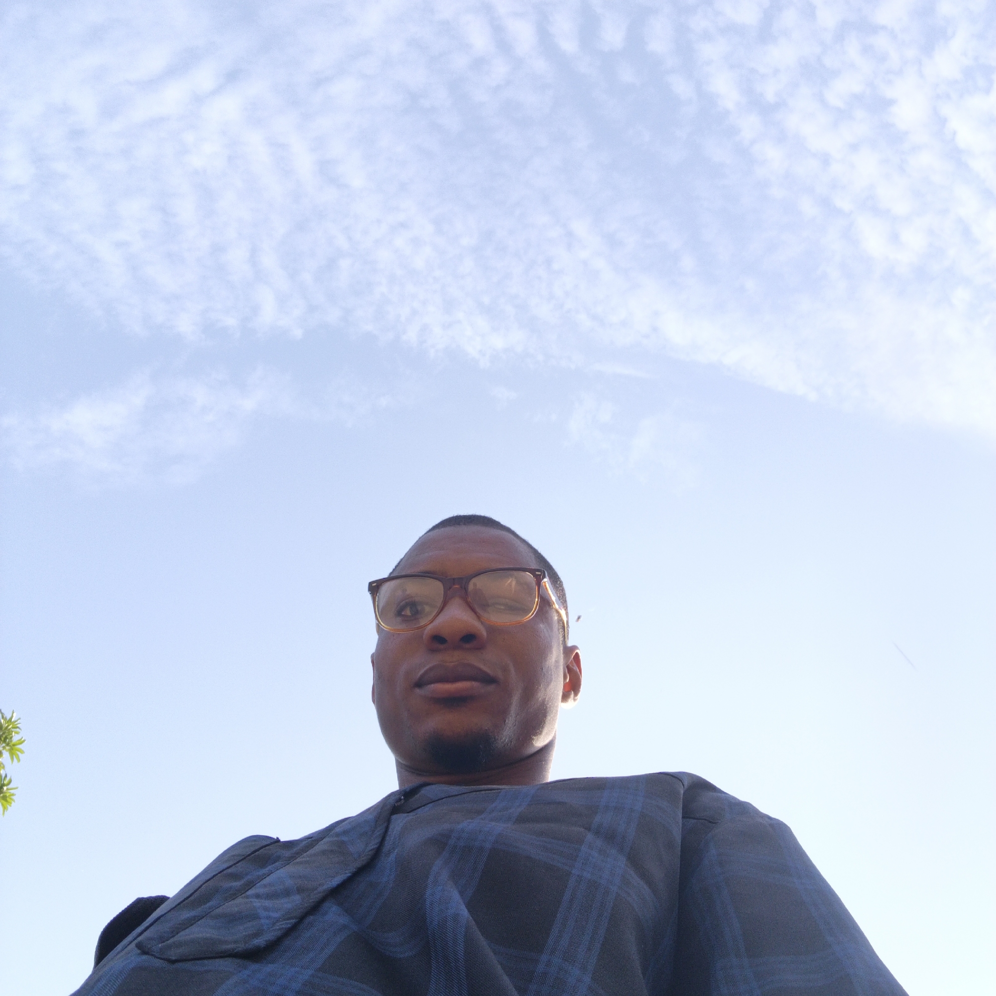 The image is a low-angle, outdoor, wide shot of a Black man wearing glasses and a blue and black plaid shirt. The man is positioned in the lower half of the frame, looking up towards the camera. His face is partially obscured by the angle, but his glasses are clearly visible. The background is a bright, pale blue sky with wispy white clouds. There is some green foliage visible on the left side of the frame, and the man's clothing suggests a warm climate. The image appears to be a selfie, taken from a low perspective. The specific location in Shaba, Nigeria is not discernible from the visual information in the image itself.