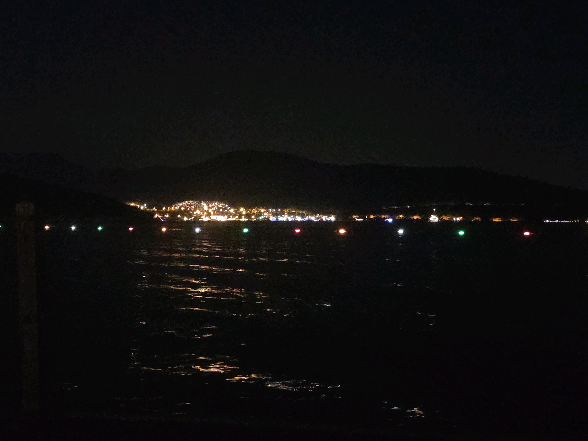 An aerial nighttime long shot captures the city of Bodrum, Turkey, illuminated by a myriad of lights that twinkle and reflect on the dark, rippling water. The distant shore is densely packed with buildings, their windows and streetlights forming a bright, continuous band of light. Above the city, the dark silhouette of a mountain range looms against the night sky. In the foreground, a series of small, colored lights—white, green, red, and yellow—are spaced out across the water, likely marking a channel or path. The surface of the water is textured with small waves, creating streaks of light and shadow. A dark, vertical object, possibly a wooden post or piling, is visible on the far left of the frame.