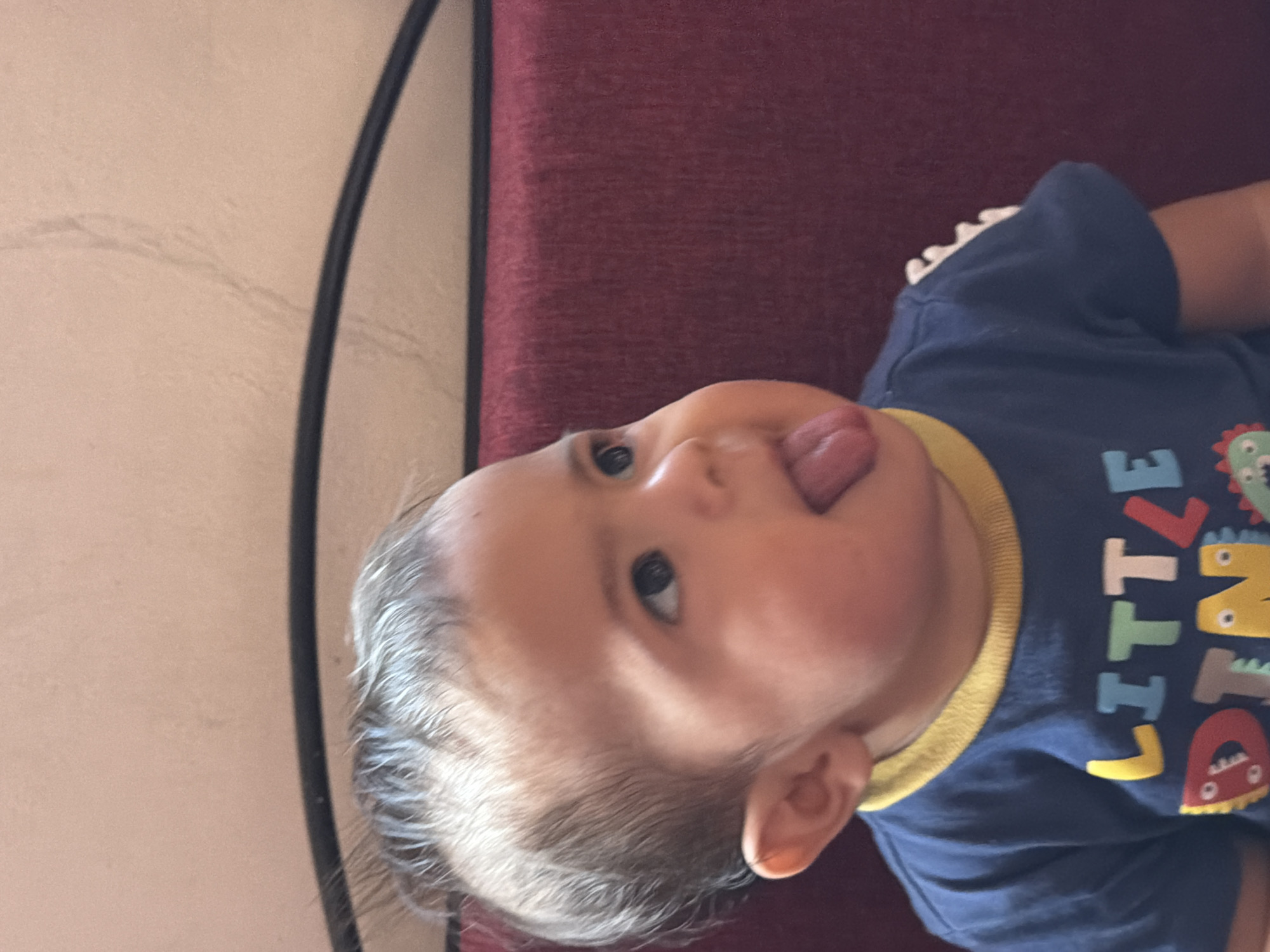 A close-up shot of a baby lying on a burgundy-colored cushion. The baby is wearing a navy blue shirt with a yellow collar and the words "LITTLE DINO" in colorful lettering. The baby's tongue is sticking out. The location is Reforma, Mexico, and the photo appears to have been taken indoors. There is a hint of a wall and frame in the background.