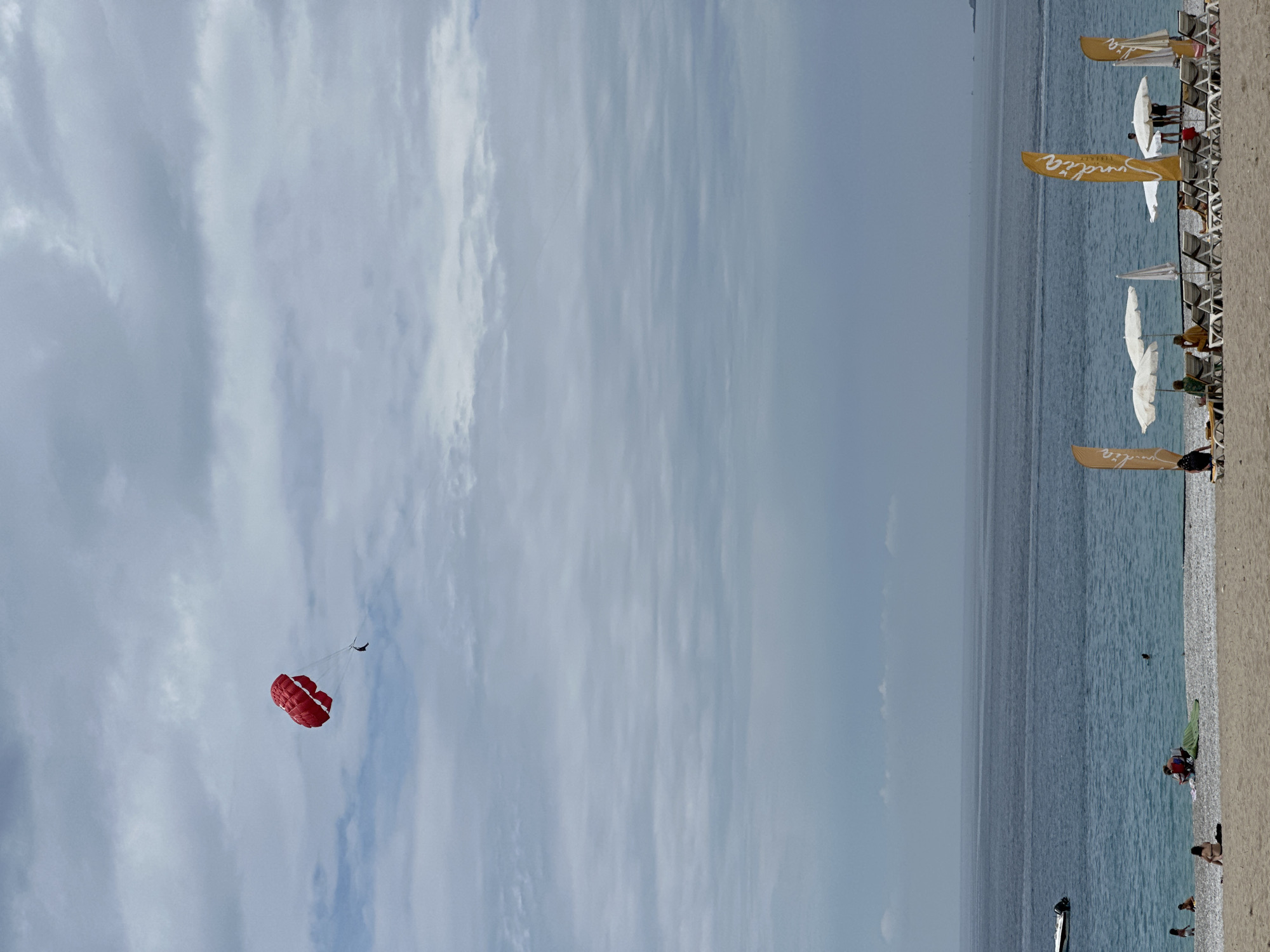 This image captures a vibrant daytime scene at a beach in Fethiye, Turkey, under a mostly cloudy sky. The horizon line clearly separates the expansive blue-grey sea from the lighter grey sky, with a strip of sandy beach running along the right side of the frame (when viewed upright).
In the upper left quadrant of the image, a bright red parasail is visible, carrying a single person high above the water. This individual appears as a small, dark silhouette suspended beneath the colorful canopy, engaged in the popular watersport.
Along the sandy beach, numerous white beach chairs and umbrellas are neatly arranged in rows, indicating a resort or organized beach area. Several people are seen relaxing on the chairs or strolling on the sand. A few individuals are also present in the calm, turquoise waters of the sea, possibly swimming or wading.
Prominently featured along the beach are several tall, golden-yellow flags. The text "Suadia RESORT" is clearly legible on these flags, providing a location cue for the specific beach establishment. The cloudy but bright sky suggests pleasant weather, ideal for leisure activities. The overall atmosphere conveyed is one of relaxation and recreation at a popular coastal destination.