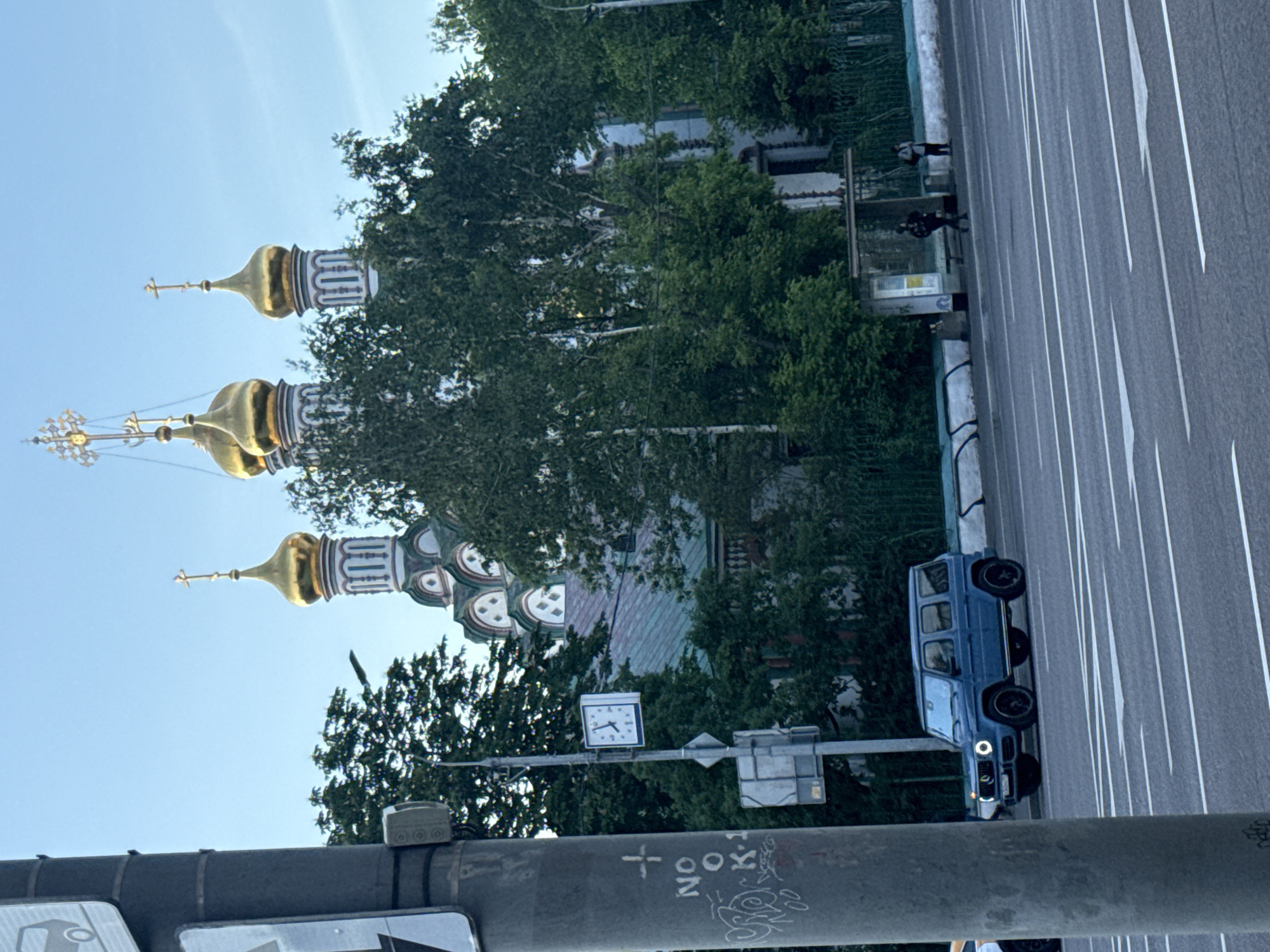 The image displays a vibrant daytime street scene in Moscow, Russia, featuring a prominent Eastern Orthodox church next to a multi-lane road.
On the left side, a grand church is visible with several golden, onion-shaped domes, each crowned with an ornate cross. The church's facade is intricately decorated with white and patterned elements, partially obscured by dense green trees. One cross features an elaborate sunburst design.
To the right, a wide asphalt road with multiple lanes is evident, marked by clear white lines and directional arrows on the pavement. A dark blue, boxy SUV, resembling a Mercedes G-Wagen, is positioned on the road with its headlights illuminated. Further along the road, two individuals are seen near a bus stop shelter, appearing to be waiting.
In the foreground, a large, dark gray utility pole or beam crosses the lower part of the frame. Attached to it is a small, square clock face with Roman numerals, showing a time of approximately 9:30. The pole also bears white graffiti, including a cross symbol and the text "NOO K".
The bright blue sky indicates a clear, sunny day. The scene captures a blend of historical architecture and modern urban life.