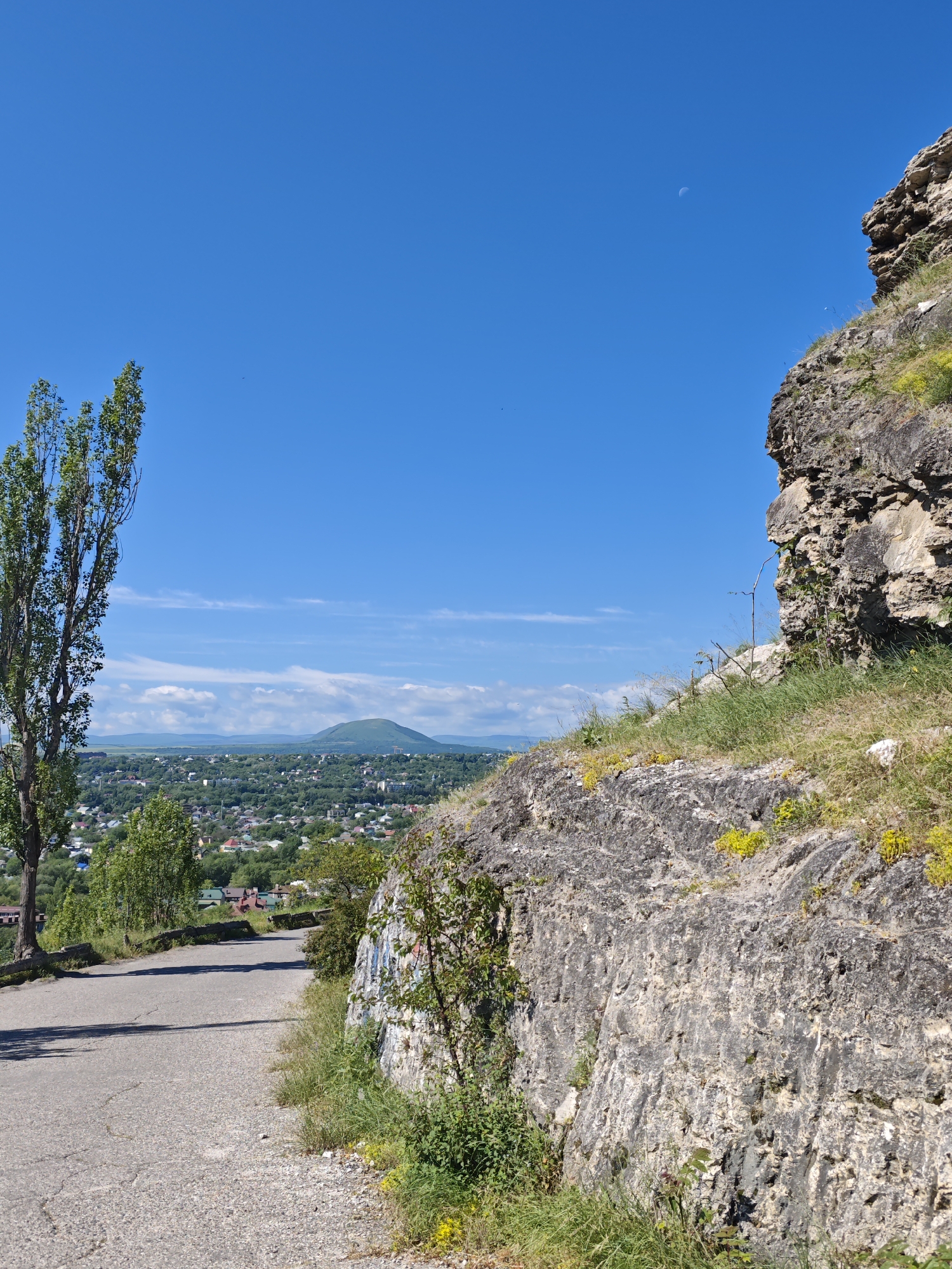 The image captures a scenic view from an elevated position overlooking the city of Goryachevodsky, Russia. In the foreground, a paved road curves upwards, bordered by a rugged, rocky embankment on the right. The embankment is sparsely covered with green grass and small yellow wildflowers. A tall, slender tree stands to the left of the road, its foliage dense and green.
Beyond the road and tree, the city of Goryachevodsky sprawls across a verdant valley. Numerous buildings with red and white roofs are nestled among lush green trees. In the distance, a prominent, conical mountain rises against a clear, bright blue sky. Wispy white clouds dot the upper portion of the sky. A faint crescent moon is visible in the upper right quadrant of the sky. The overall impression is one of a tranquil, sun-drenched day in a picturesque Russian locale.
