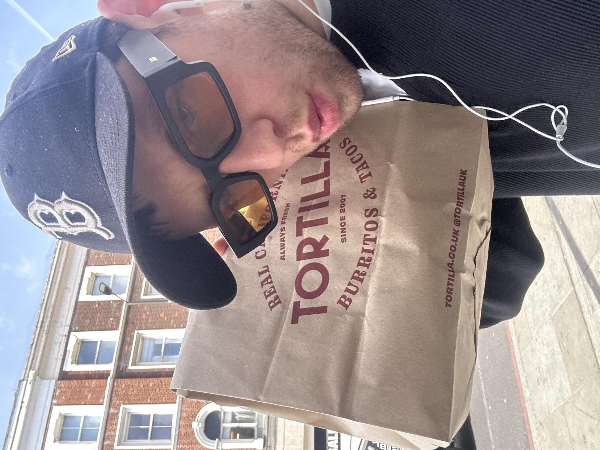 A man is photographed from a slightly angled, low-angle perspective. He is wearing a dark baseball cap with a logo on the front, sunglasses with thick black frames, and earphones. The man is holding a brown paper bag from "Tortilla," a food chain specializing in burritos and tacos. The bag's text includes "REAL CALIFORNIA," "TORTILLA," "BURRITOS & TACOS SINCE 2007," and the website "TORTILLA.CO.UK @TORTILLAAUK." The background shows a brick building with white-framed windows and a stone archway on the ground floor. The scene is likely set on a sidewalk in London, United Kingdom.