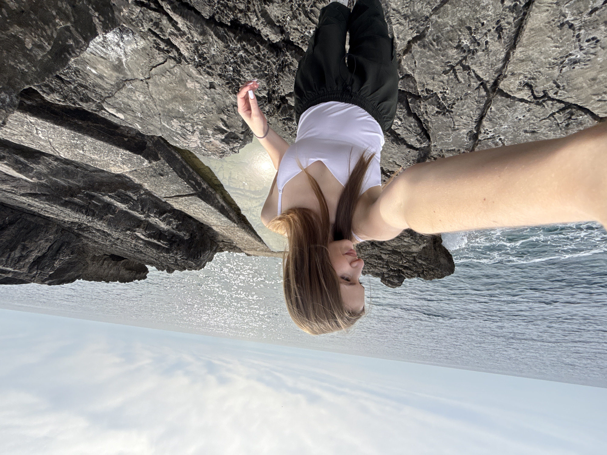 A rotated eye-level shot depicts a woman in black pants and a white tank top standing on a rocky coastline, possibly in Budva, Montenegro. The background features the Adriatic Sea meeting a cloudy sky. She appears to be taking a selfie with her right arm extended towards the camera. The rocky coastline consists of grey rocks.