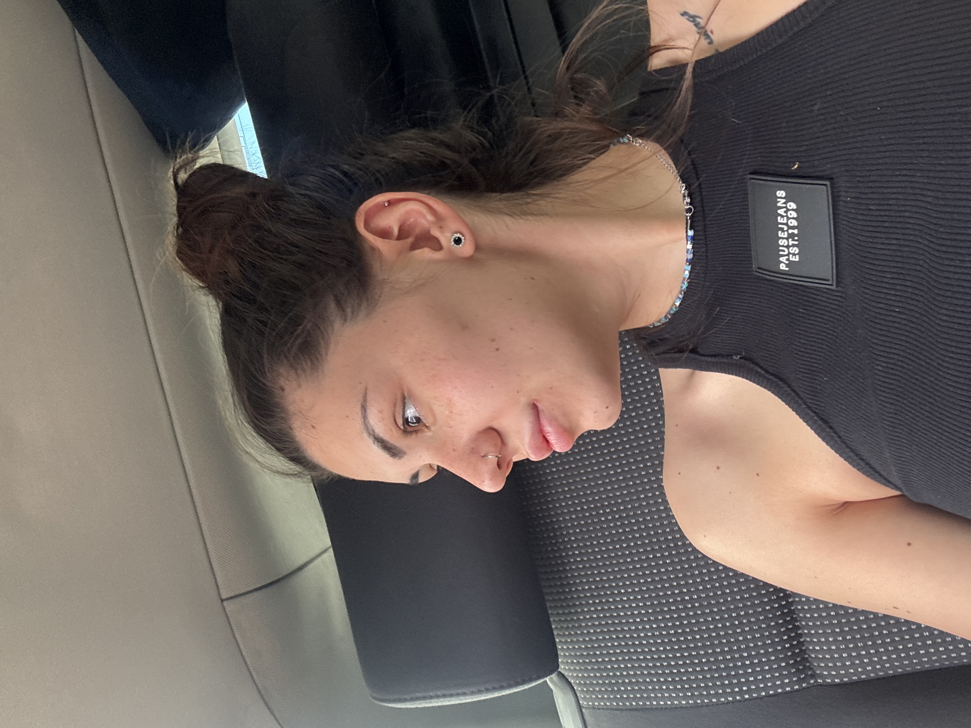 A young woman is shown in a close-up, rotated portrait, seemingly seated inside a vehicle, likely a car or bus, with the location context provided as Sofia, Bulgaria. She is looking downwards and slightly to her left (viewer's right), giving a contemplative or relaxed impression.
She has fair skin dotted with numerous freckles, particularly across her nose and cheeks. Her dark hair is pulled back into a messy bun or ponytail. Her left ear features multiple piercings, including a prominent black and silver stud in the lobe and a smaller silver stud higher on the helix. A small silver hoop nose ring is visible on her right nostril.
She is wearing a fitted, ribbed black tank top or camisole. On the upper right side of her top (viewer's left), a black rectangular patch with white text reads "PAUSE JEANS EST. 1999". A delicate necklace, composed of a silver chain and small, iridescent blue and silver beads, adorns her neck. A small, dark tattoo is partially visible on her collarbone area.
The background shows the interior of the vehicle. She is leaning against a dark grey car seat fabric with a subtle white dotted pattern. A black headrest is positioned behind her. The upper part of the vehicle's interior, possibly a pillar or roof lining, is a light beige or grey color. The lighting suggests it is daytime. No specific activity is depicted beyond her resting or observing her surroundings.